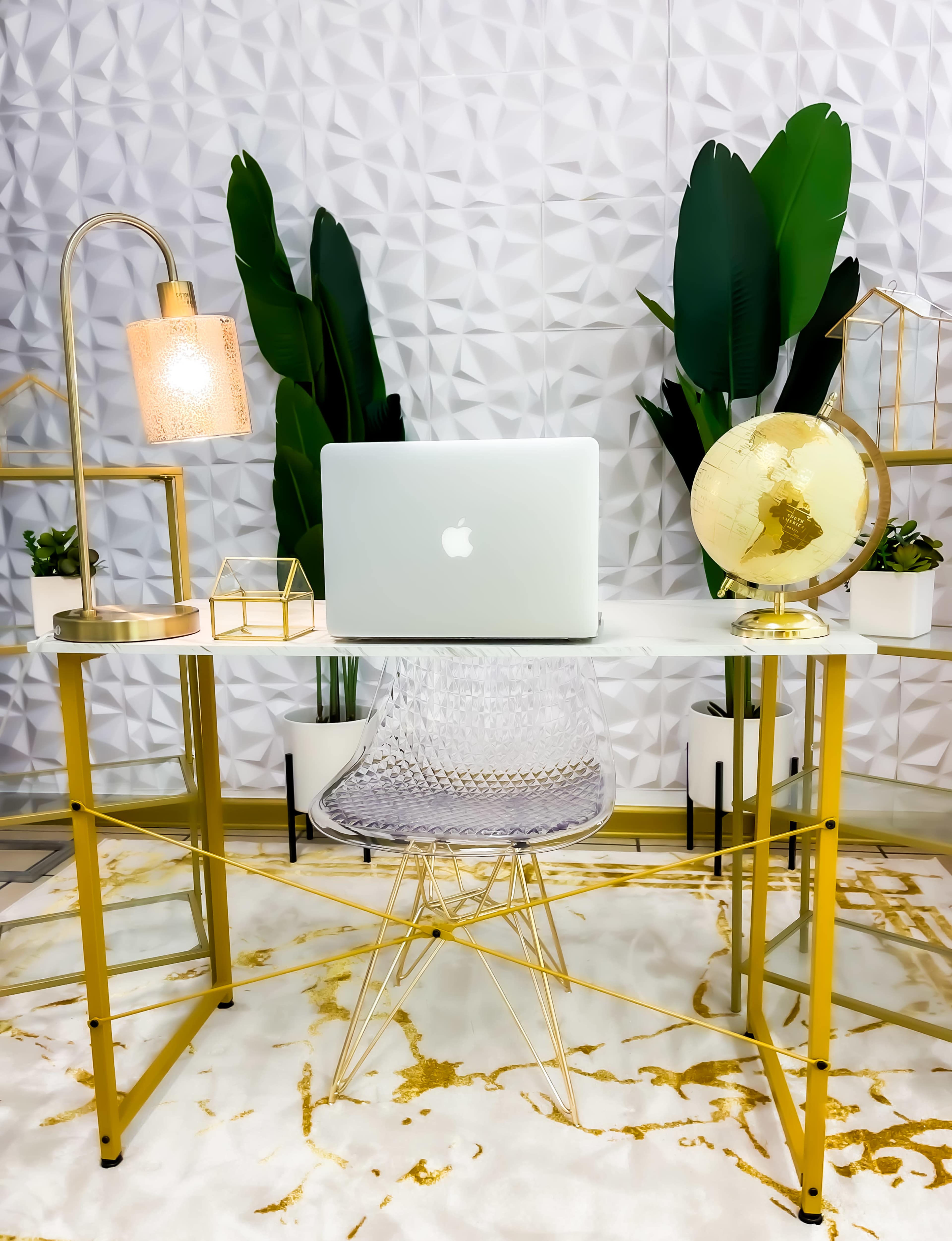 A modern workspace features a marble table with a laptop, a lamp, a globe, and potted plants against a textured wall.