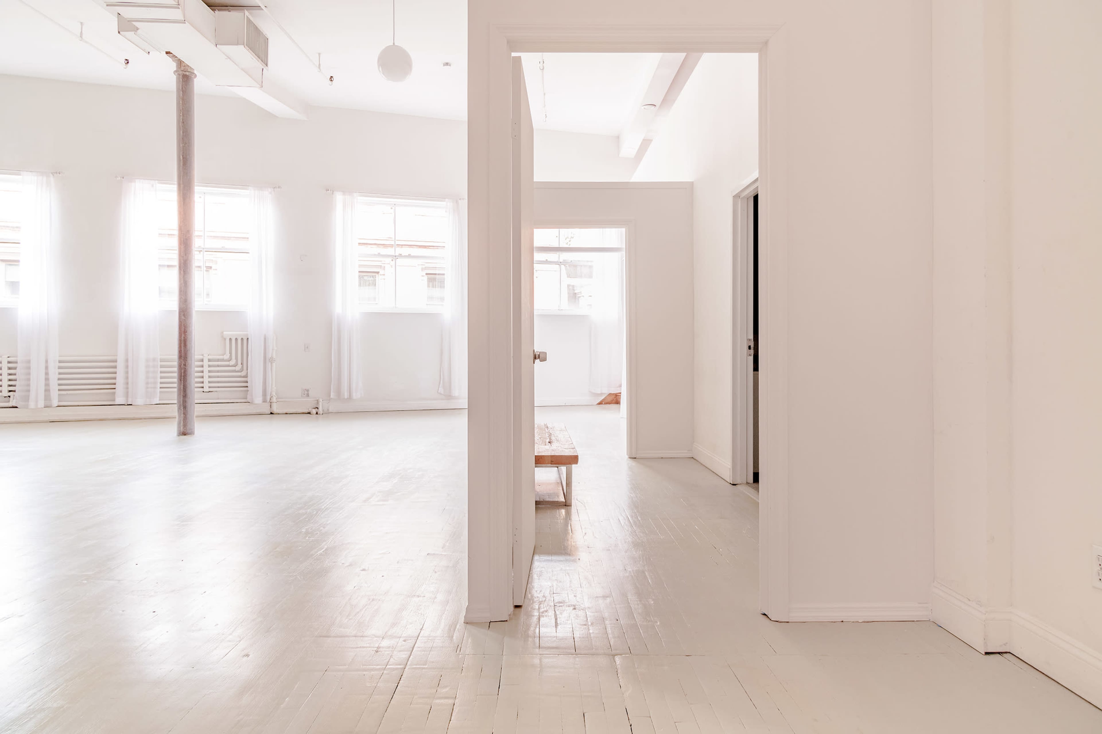 A minimalist, brightly lit interior space with light-colored wooden floors and white walls, featuring doorways leading to additional rooms.