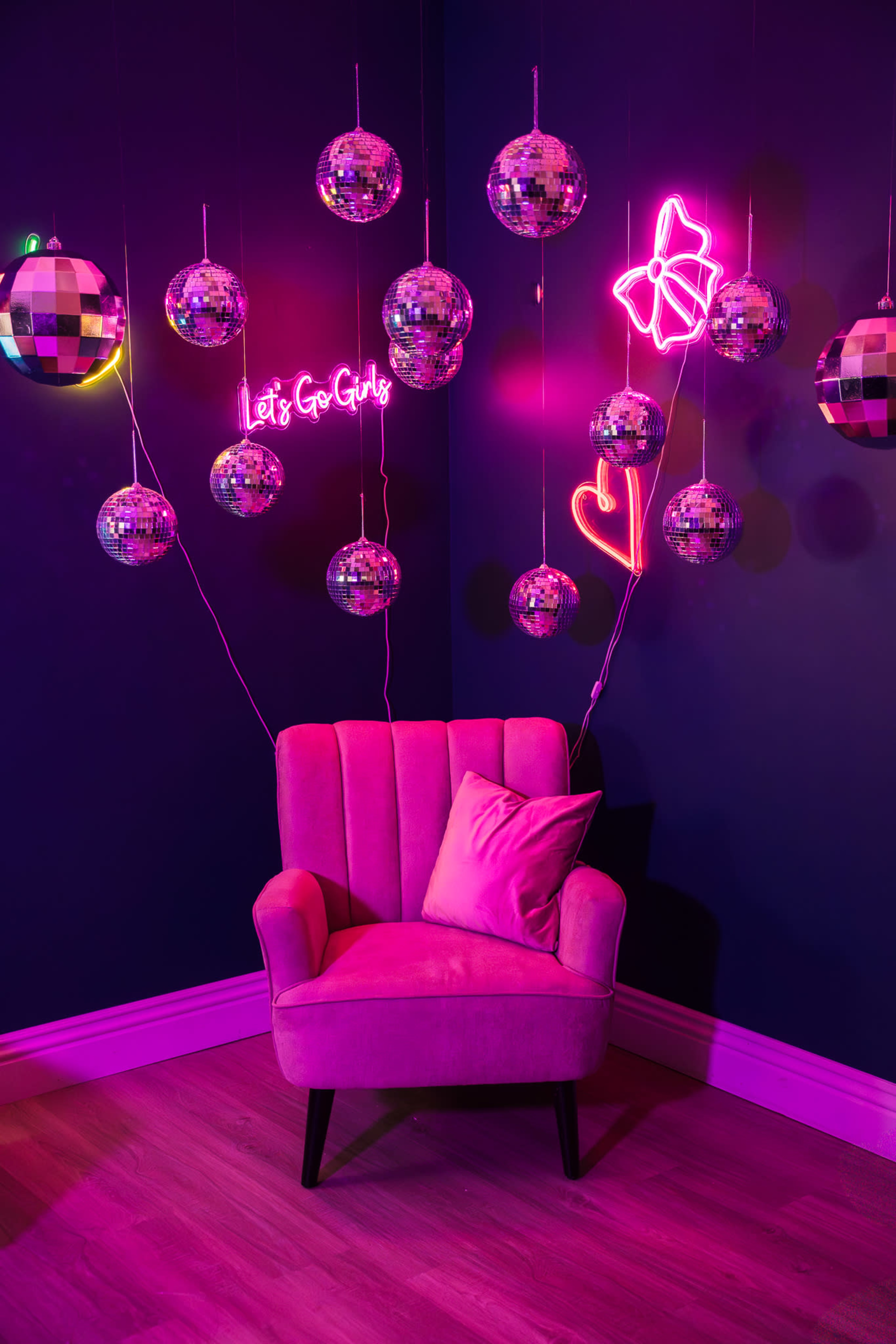 A pink velvet chair is positioned against a dark blue wall decorated with hanging disco balls and neon signs.