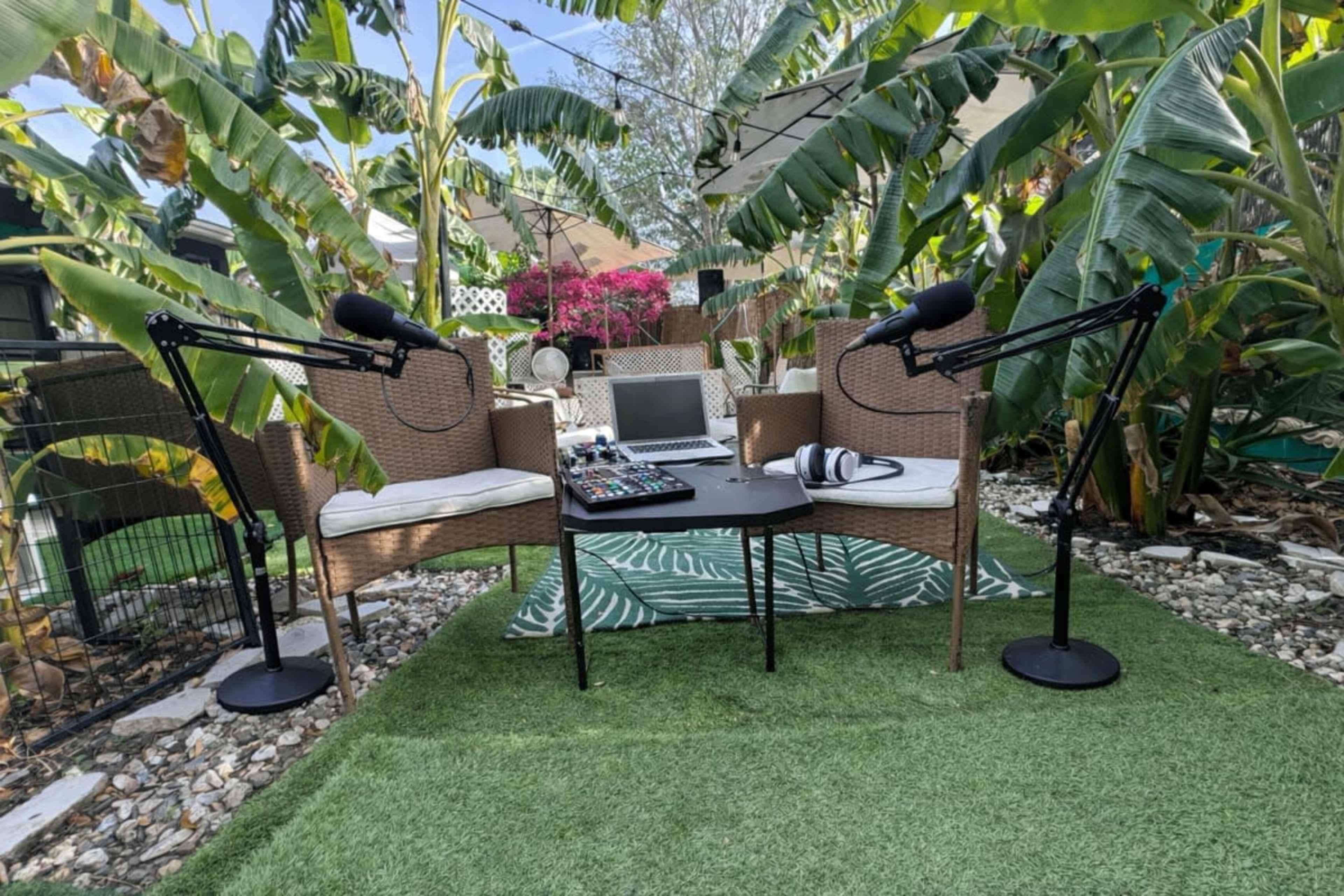 The image shows an outdoor podcasting setup with two chairs, microphones, a mixing board, and greenery surrounding the area.
