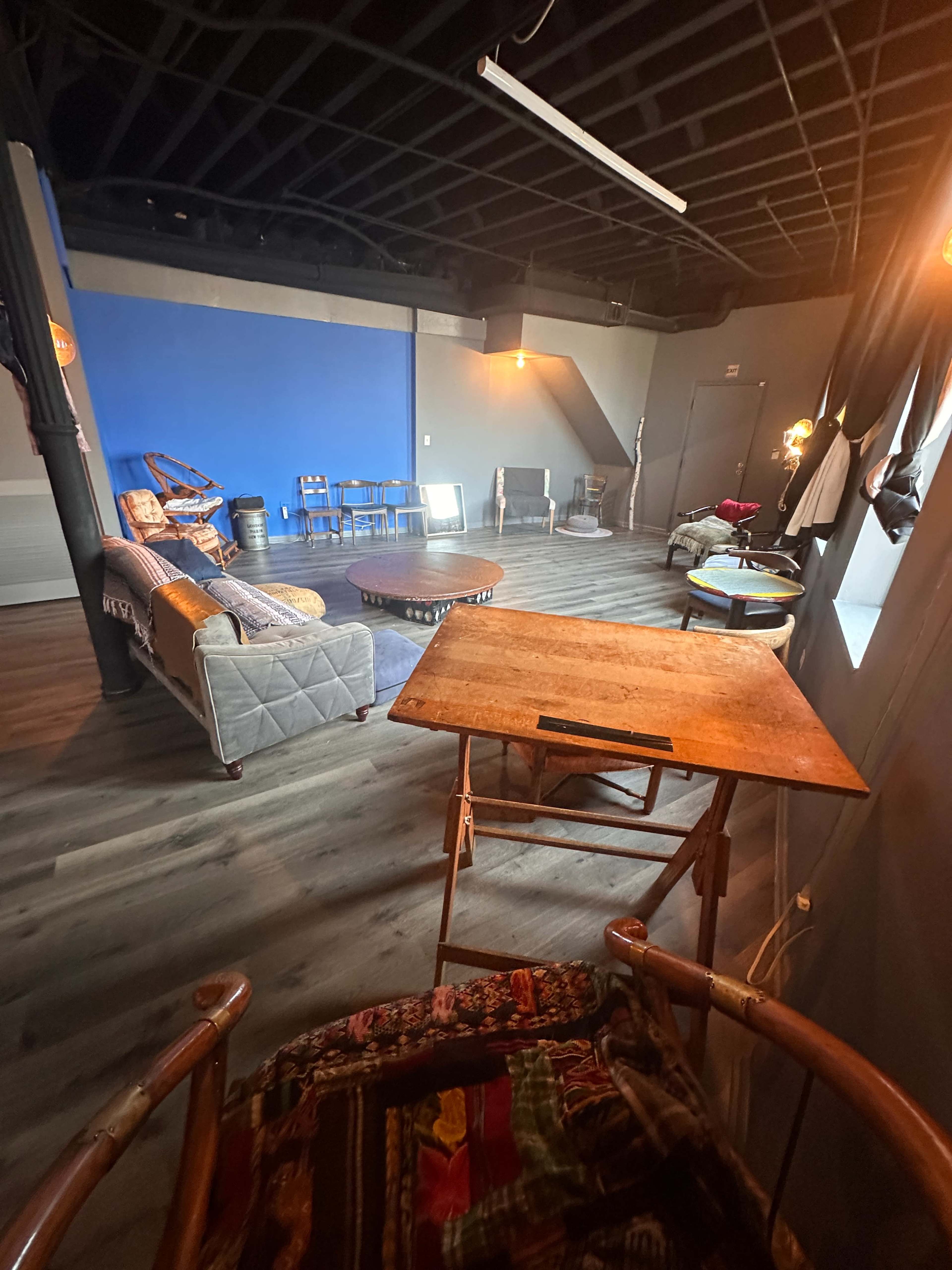 The image shows a spacious, dimly lit room with wooden flooring, featuring various seating arrangements, a coffee table, and a blue accent wall.
