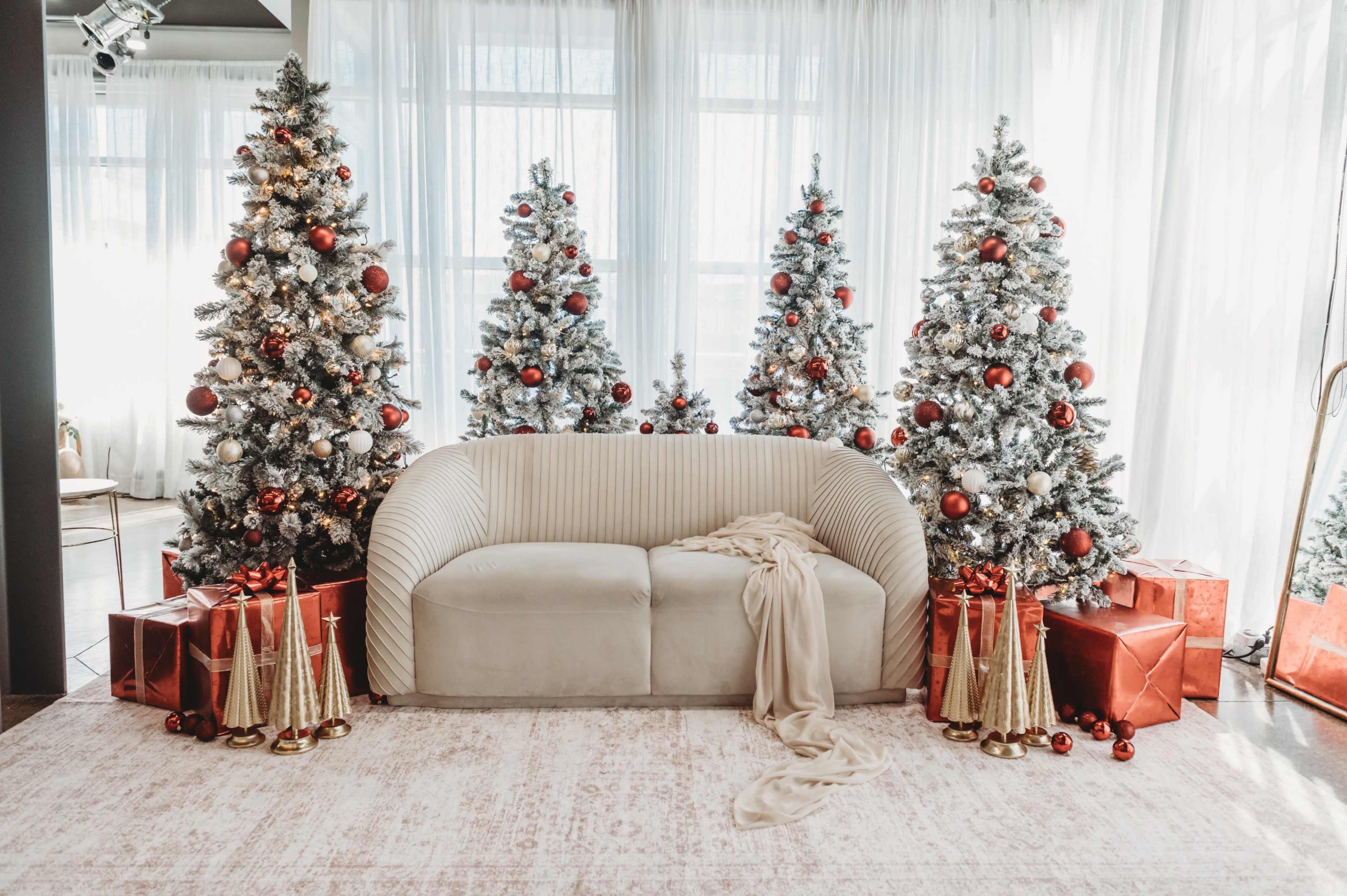 A modern living room is decorated for the holidays with a plush beige sofa, three decorated Christmas trees, and gold and red gift boxes.