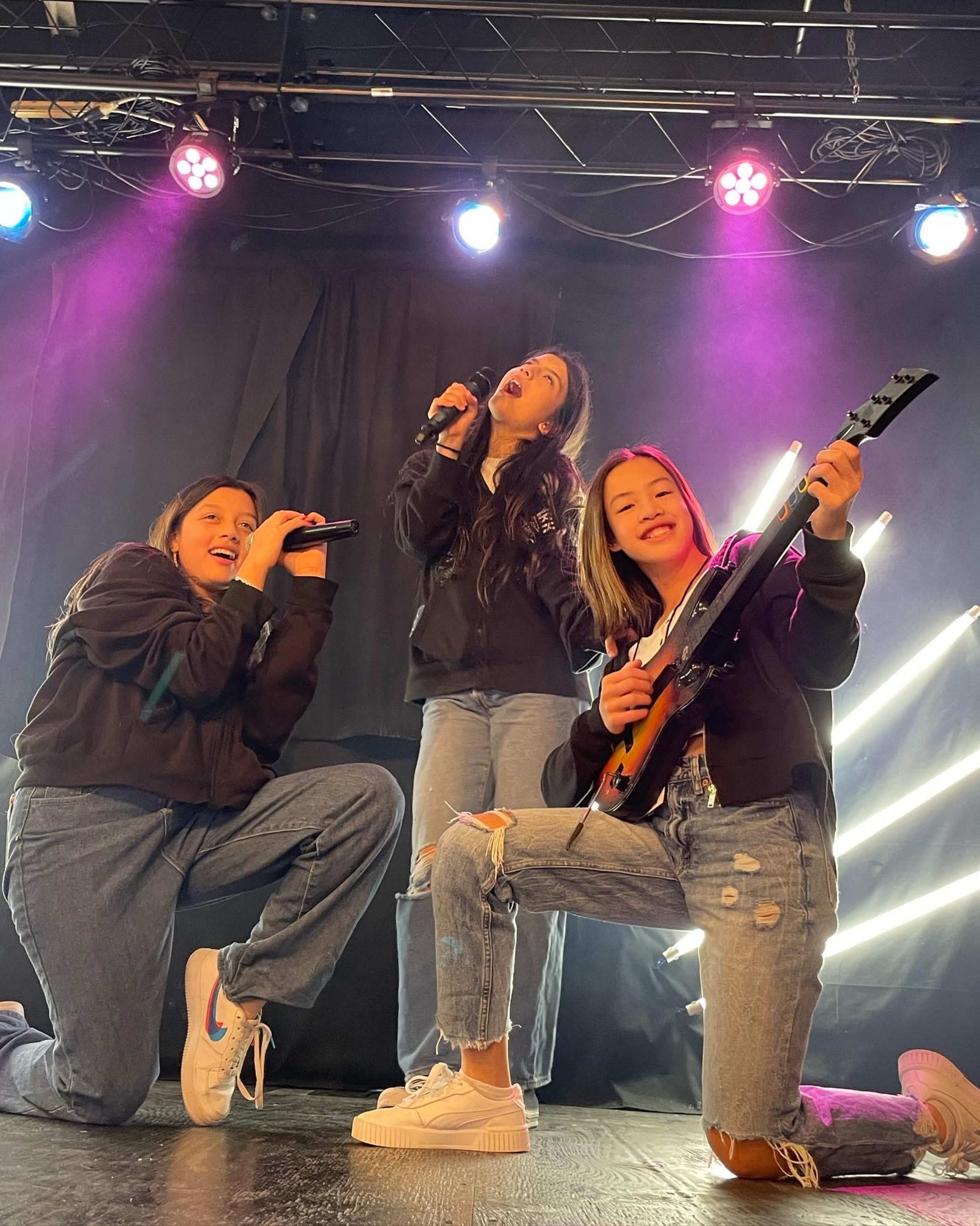 Three individuals perform energetically on stage, with one singing into a microphone and the other two joining with a guitar and microphone, illuminated by colorful stage lights.