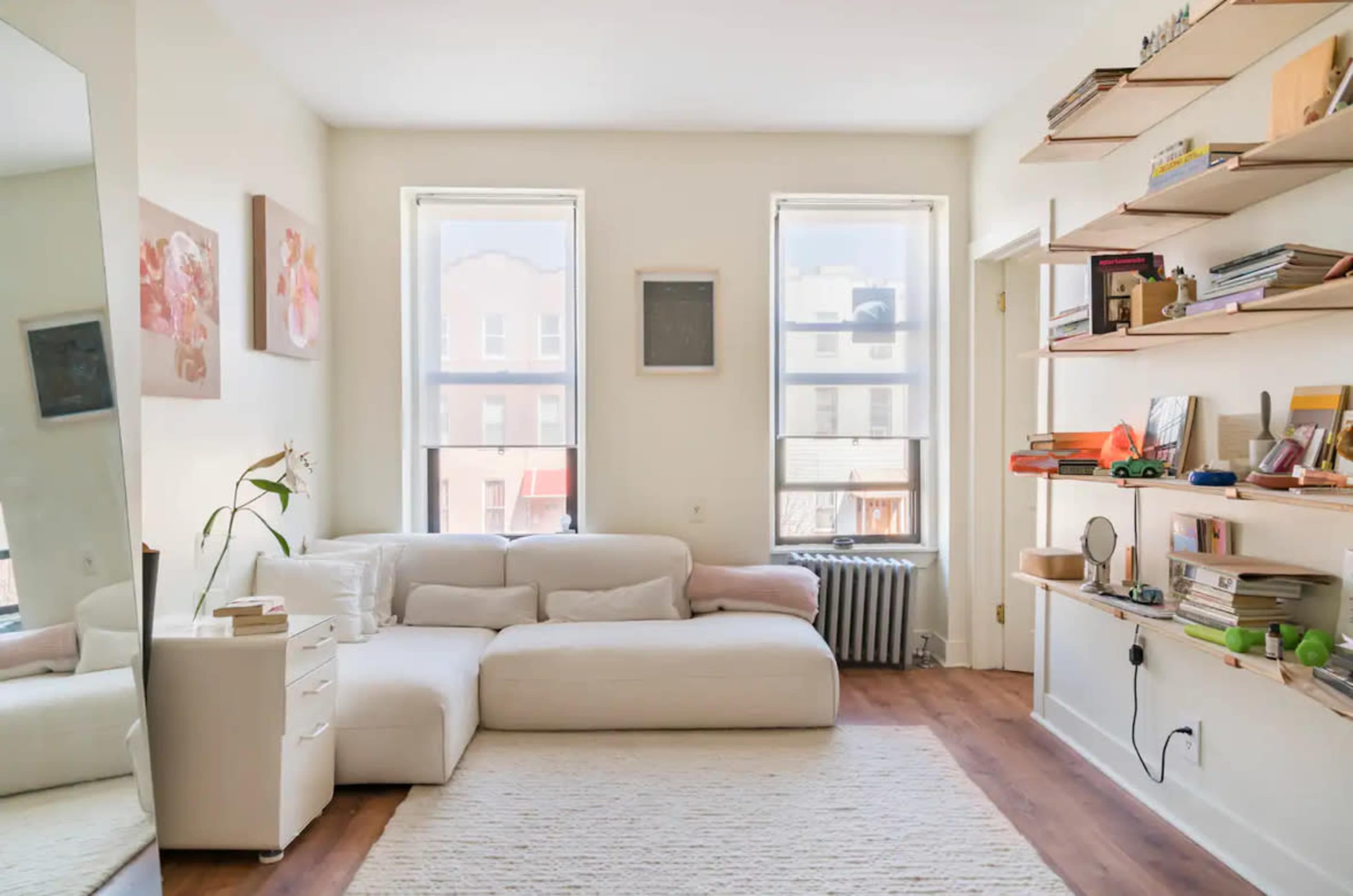 The image shows a bright, modern living room with a white sectional sofa, wooden shelves filled with books and decorative items, and large windows letting in natural light.