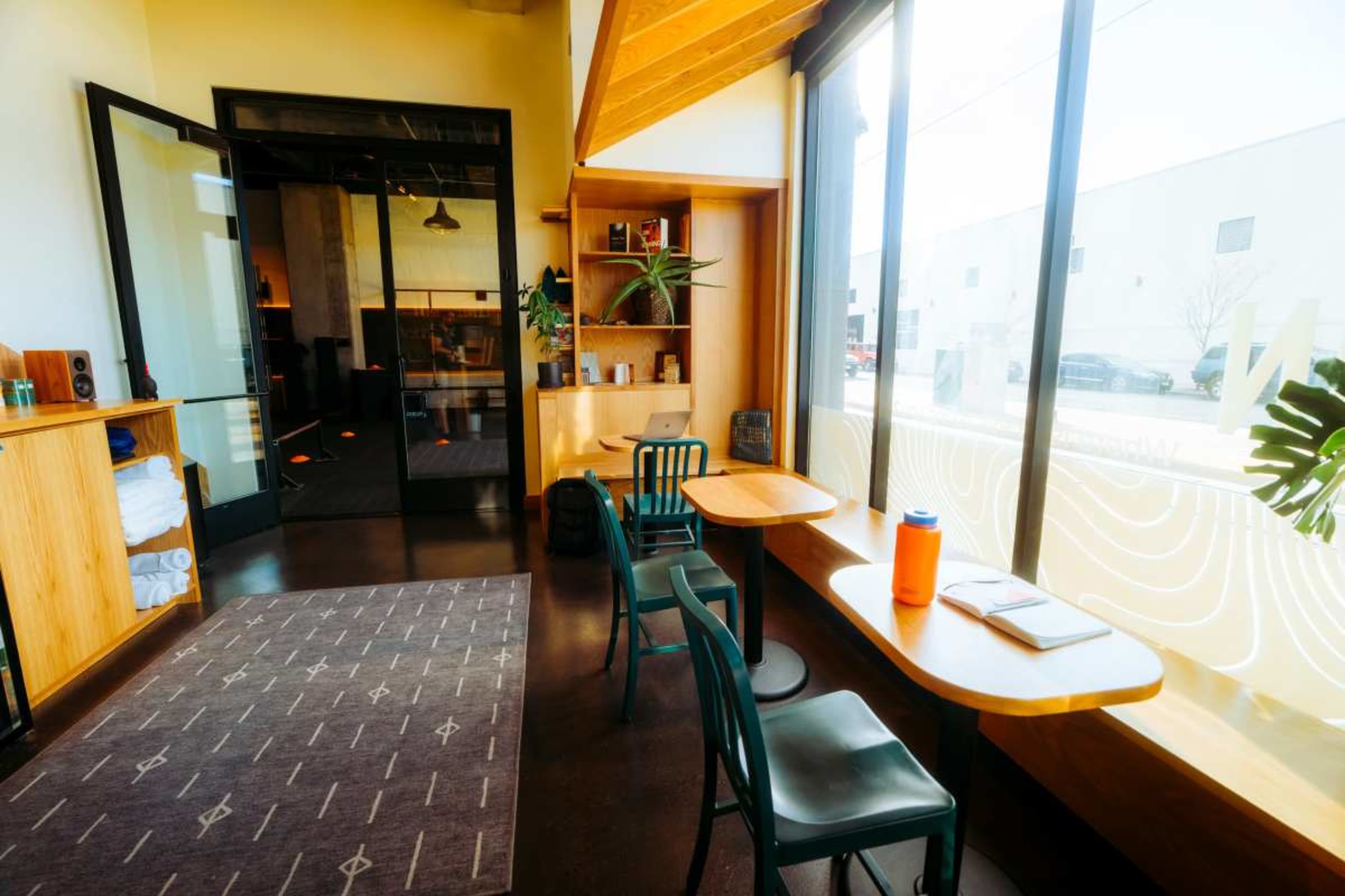 The image shows a modern café interior with two wooden tables and green chairs, large windows allowing natural light, and a bookshelf in the corner.