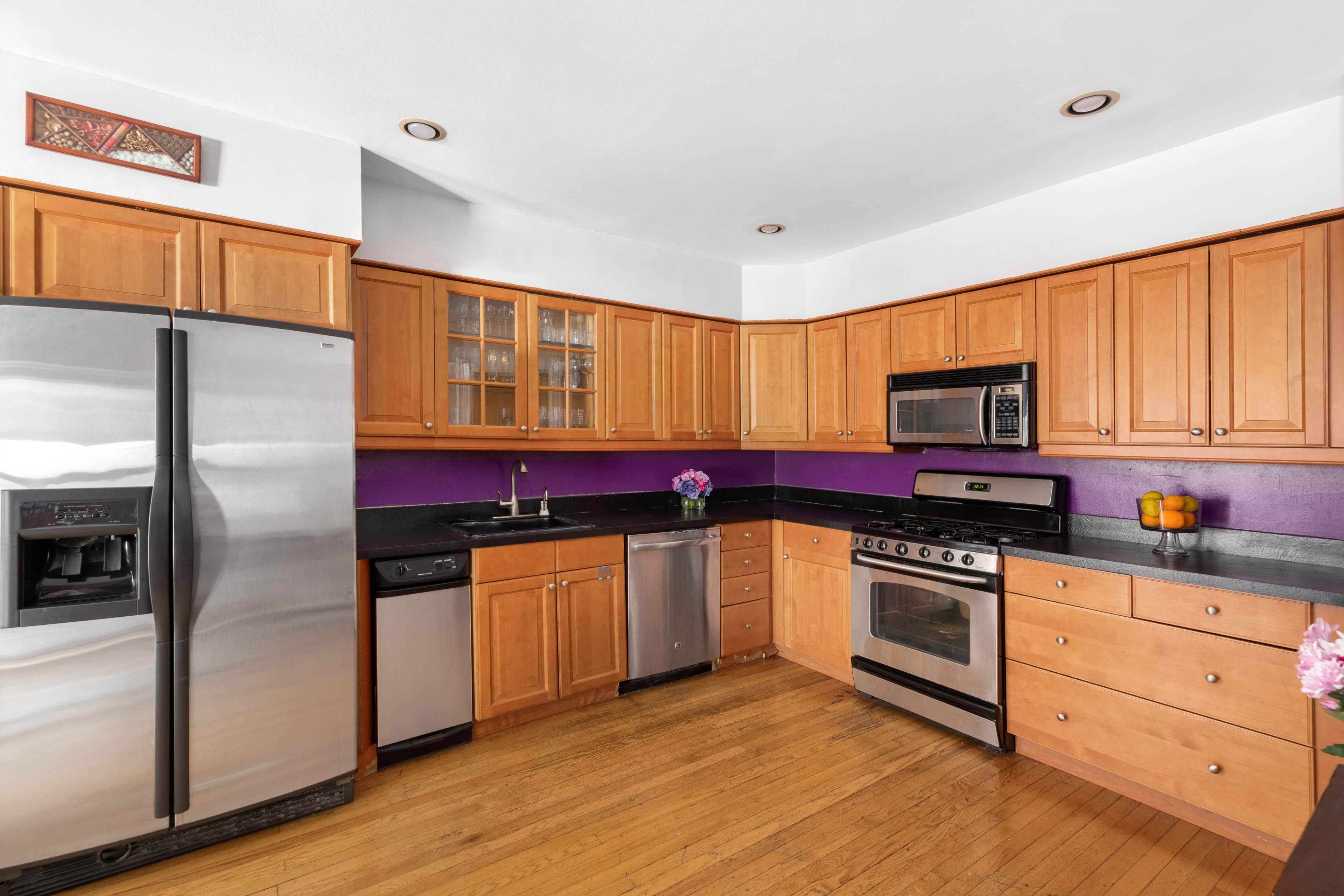 A kitchen with wooden cabinets, stainless steel appliances, a black countertop, and a purple accent wall.