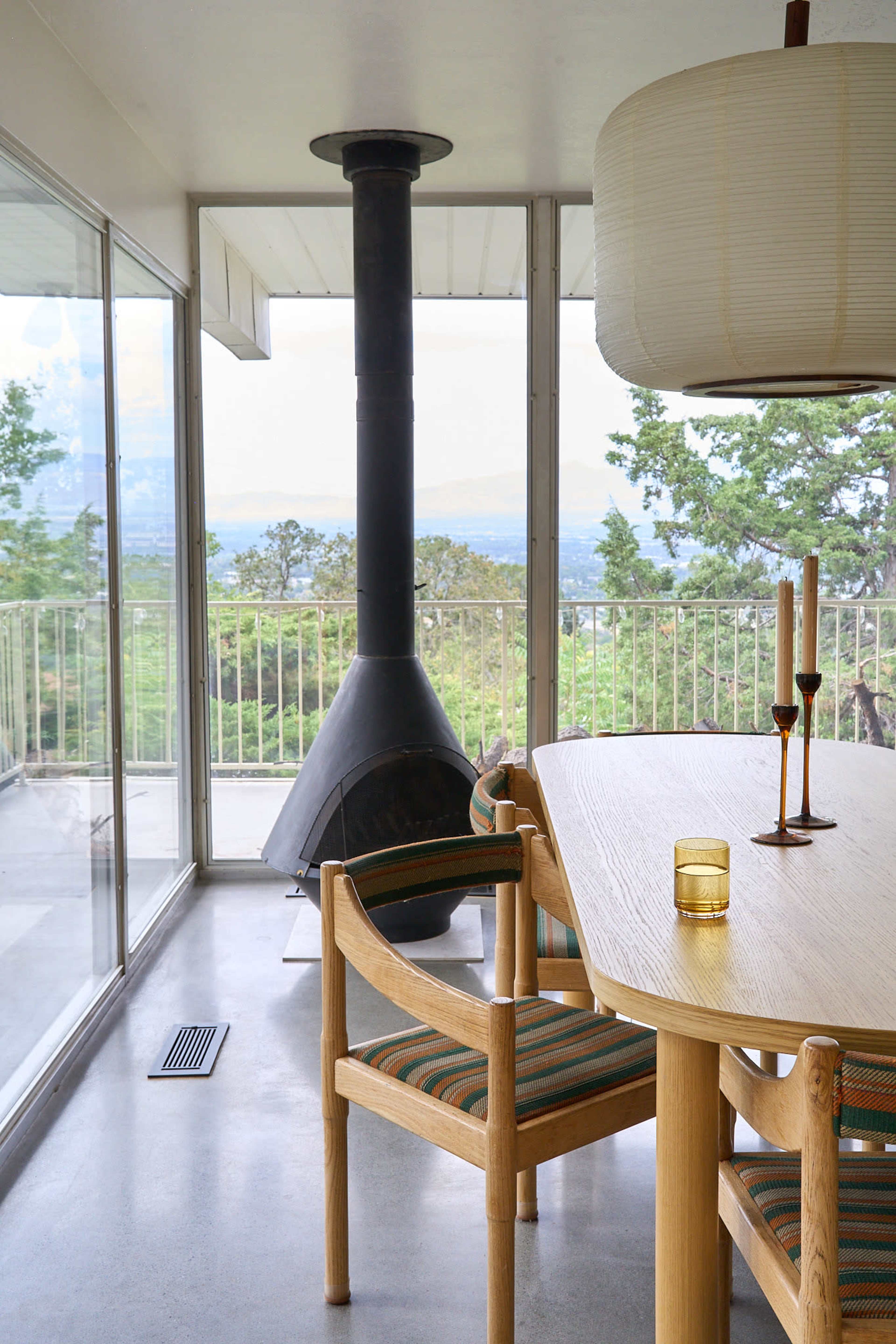 A modern dining area features a wooden table, chairs with padded seats, and a central black chimney stove, all set against large windows overlooking a green landscape.