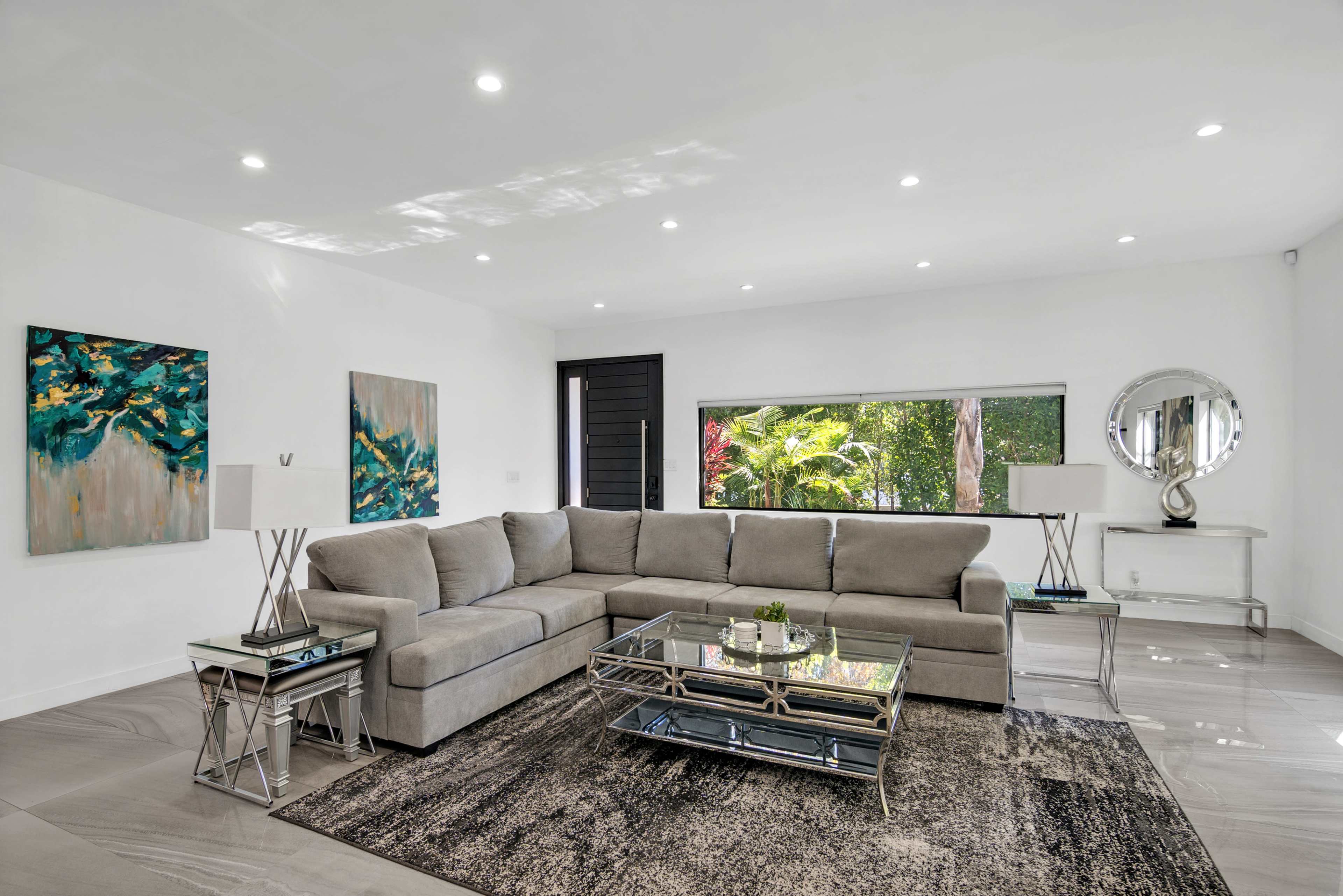 A modern living room features a large gray sectional sofa, a glass coffee table, and two lamps, with abstract paintings on the walls and a window overlooking greenery.