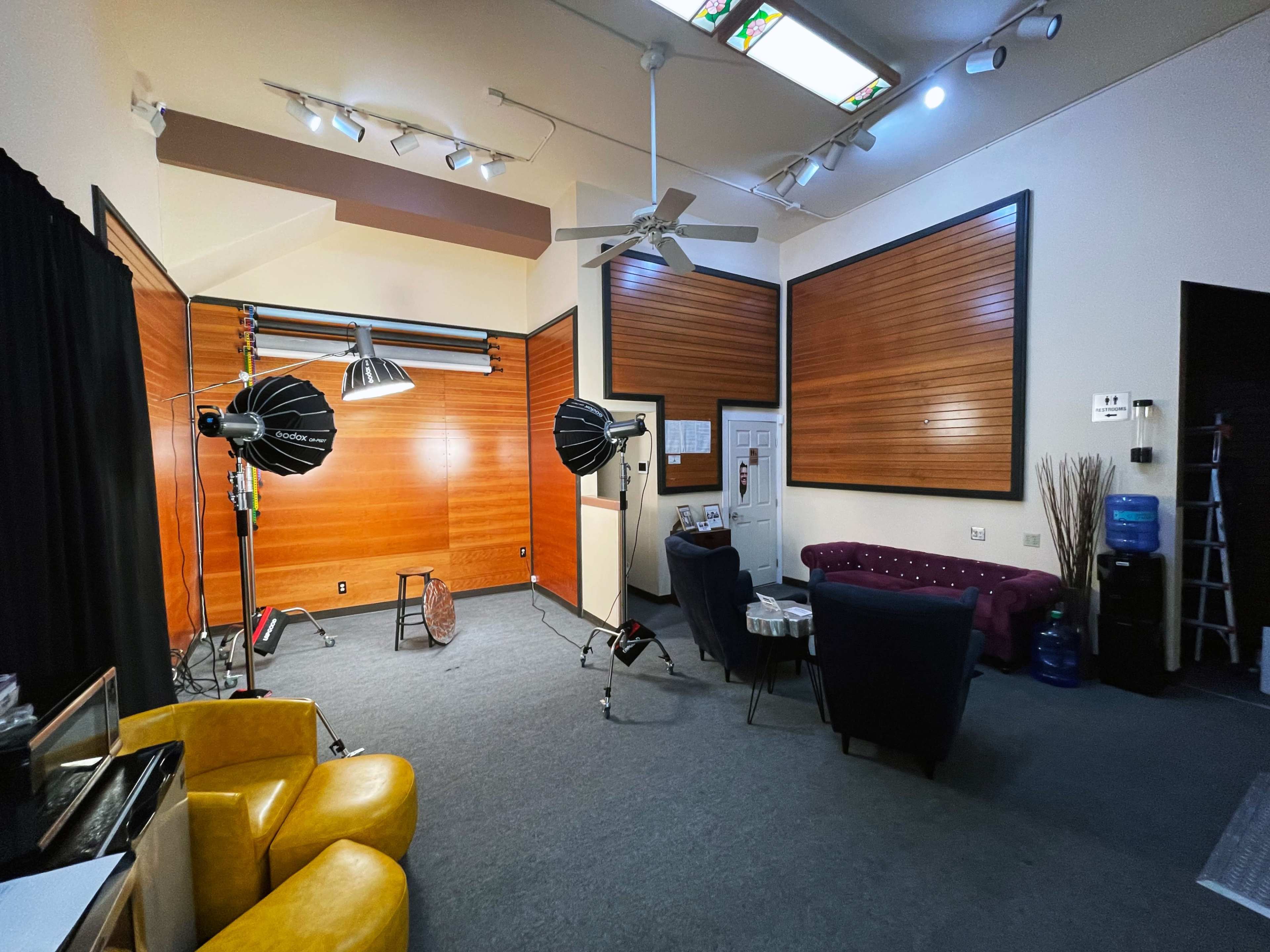 A spacious indoor room with wooden paneling, featuring two studio light setups, a burgundy sofa, black chairs, and a ladder in one corner.
