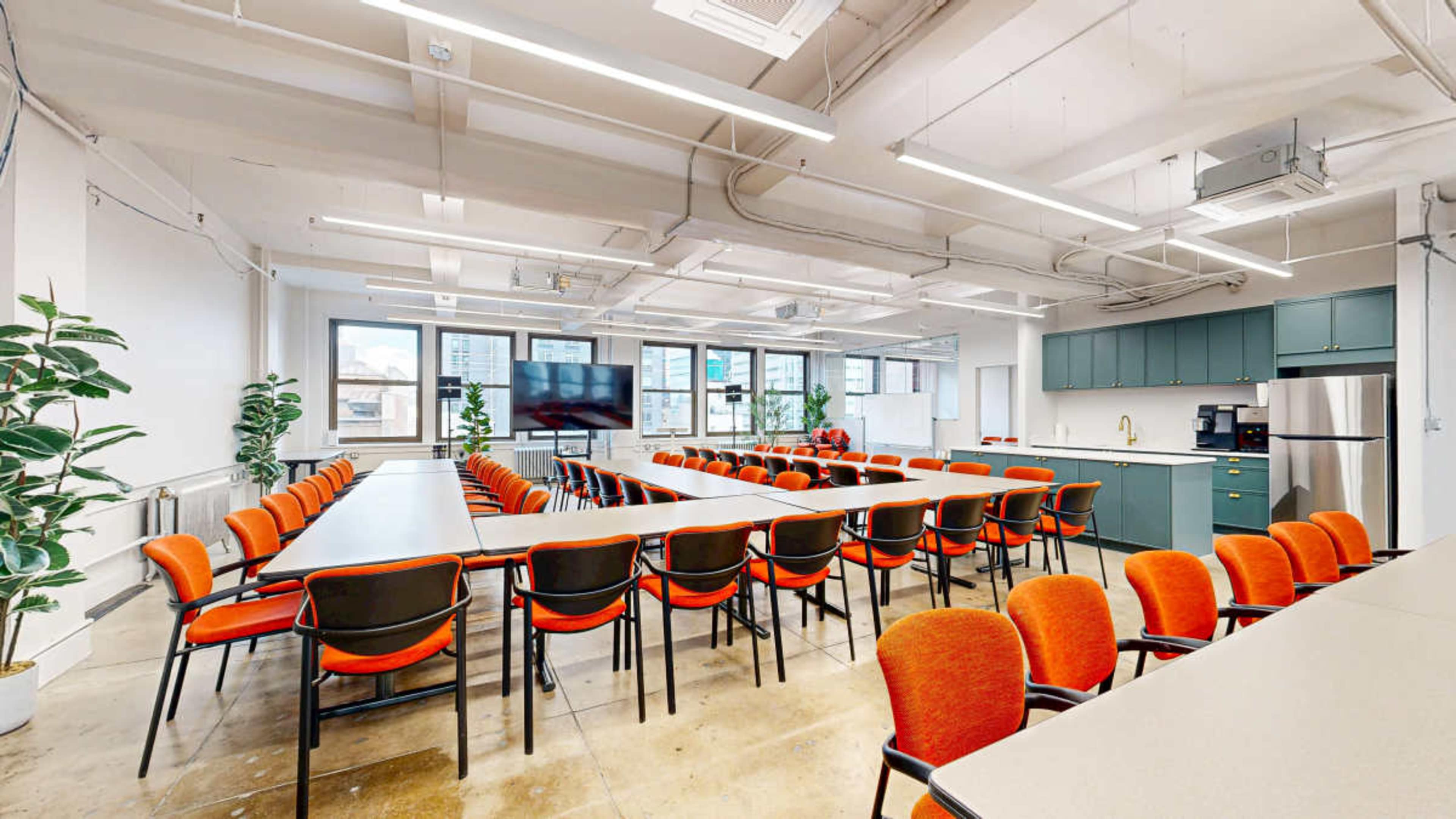 The image shows a modern conference room with a large, rectangular table surrounded by orange chairs, and large windows providing ample natural light.