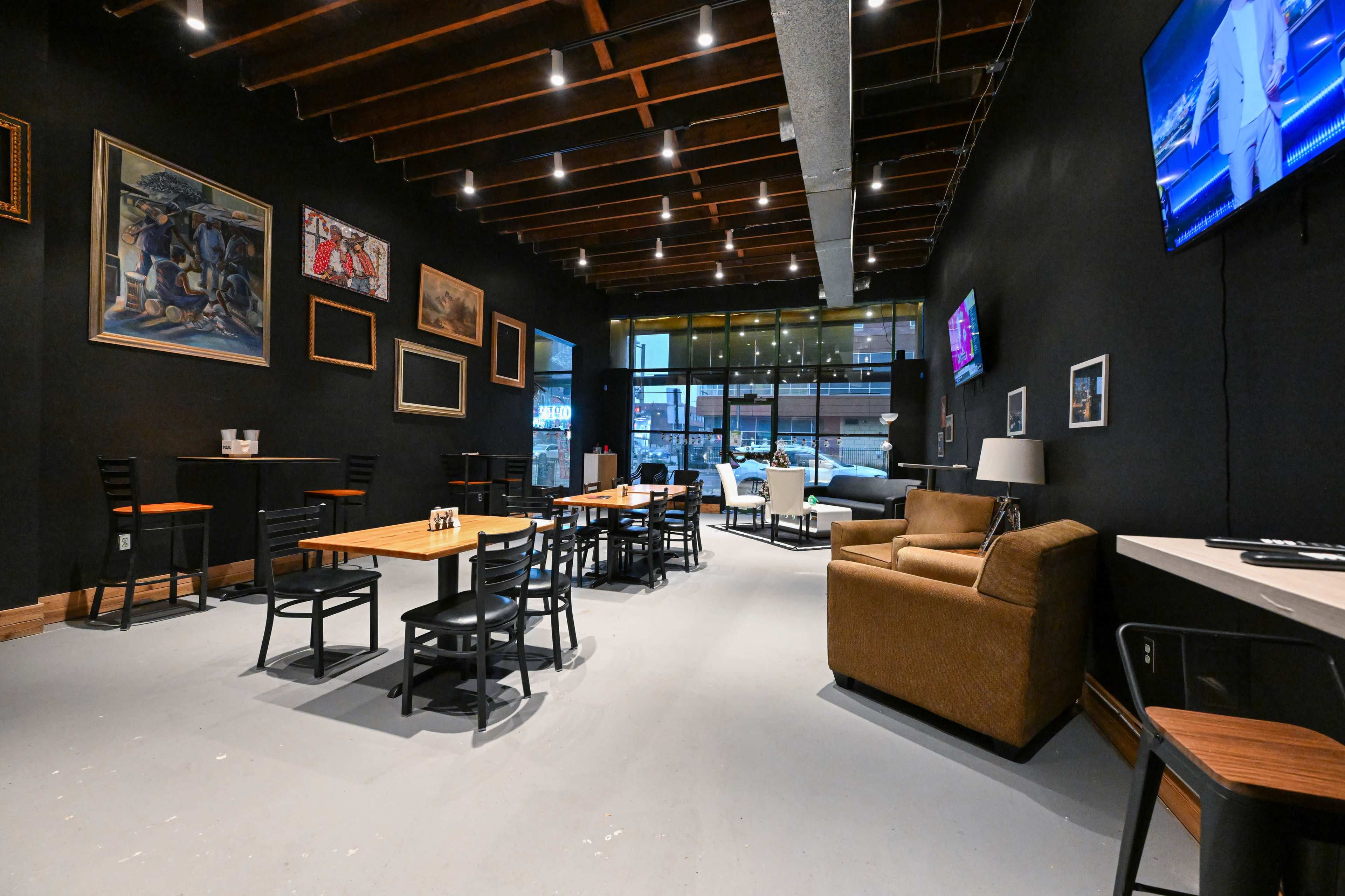 The image shows the interior of a spacious restaurant with black walls, wooden beams, various artworks, and seating arrangements including tables, chairs, and a sofa.