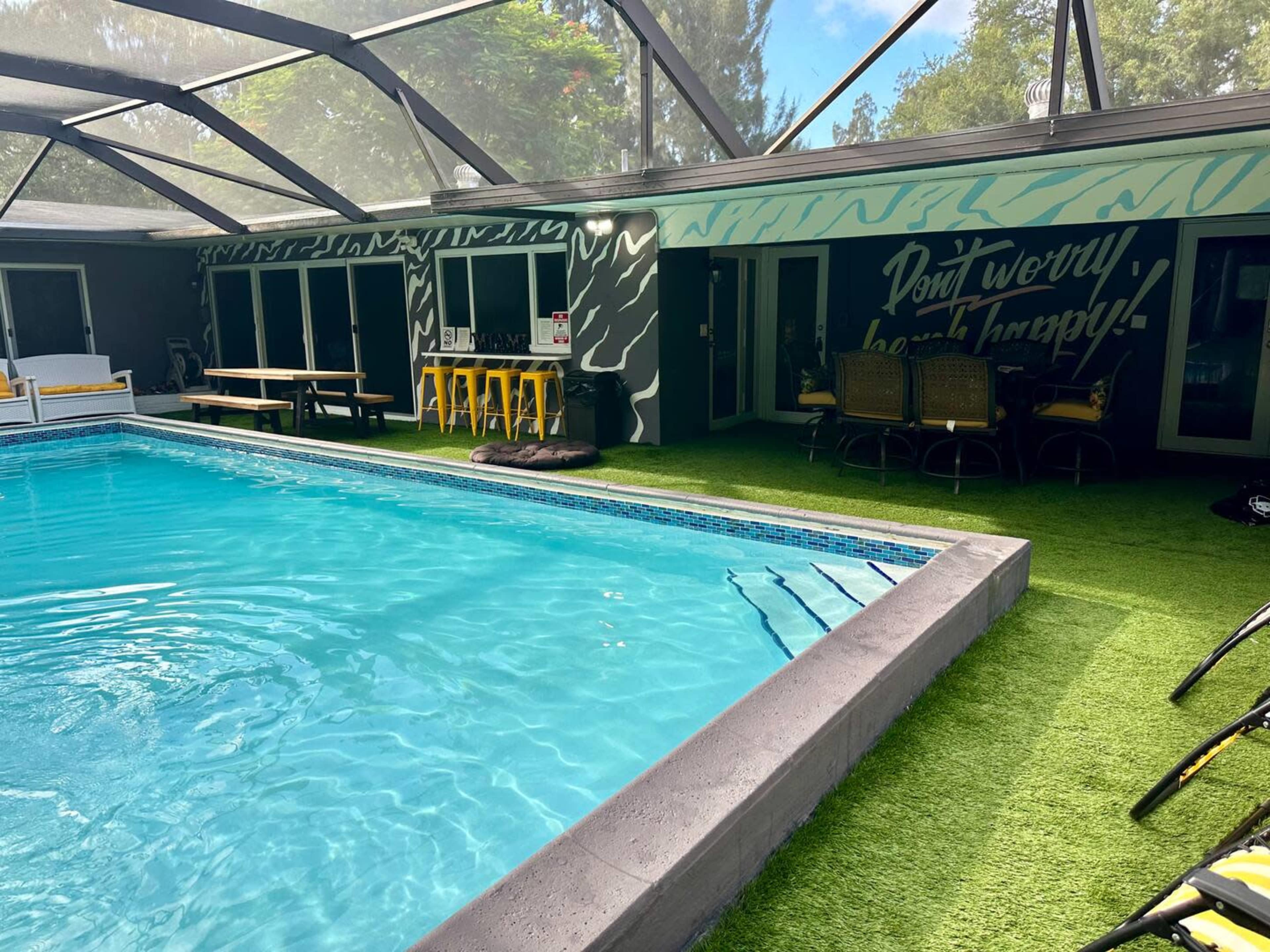 The image shows a swimming pool surrounded by a glass enclosure, with seating areas and a colorful mural on the wall.