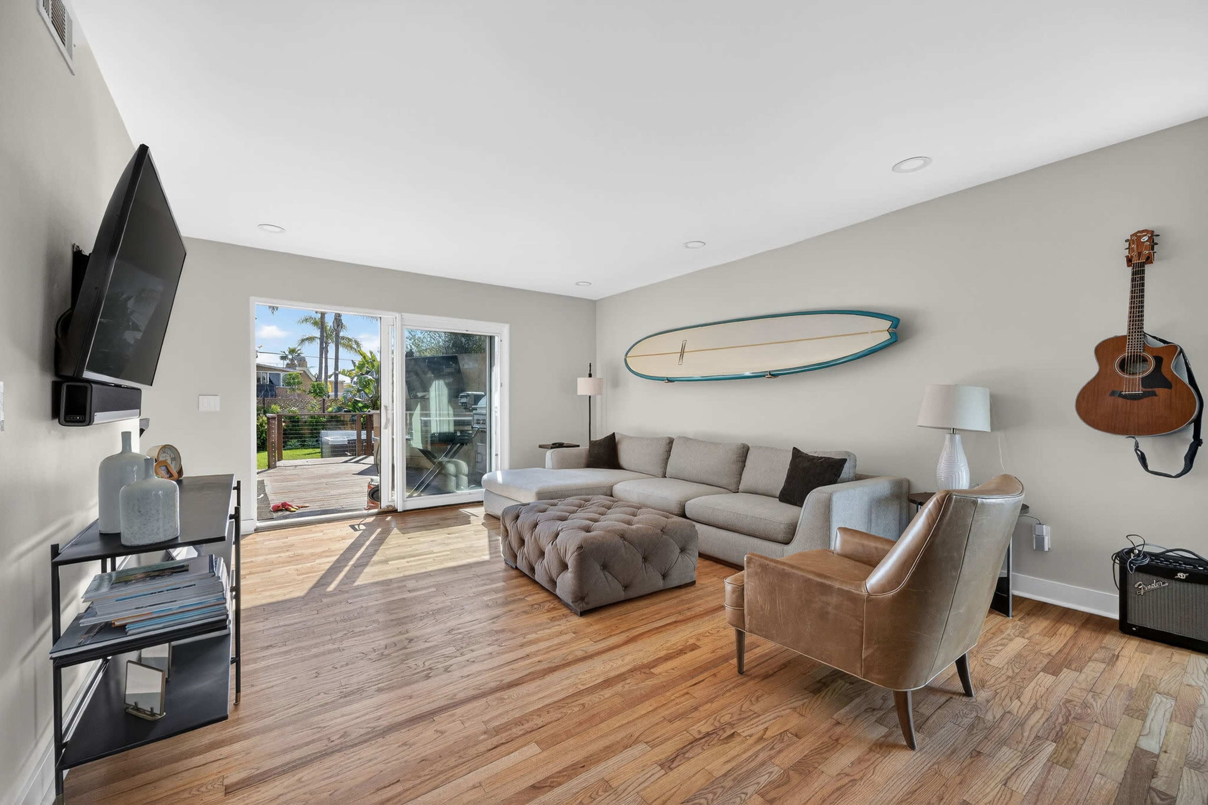 A light-filled living room features a gray sectional sofa, a tufted ottoman, a wooden surfboard mounted on the wall, and an acoustic guitar hanging next to a lamp and a black shelving unit.