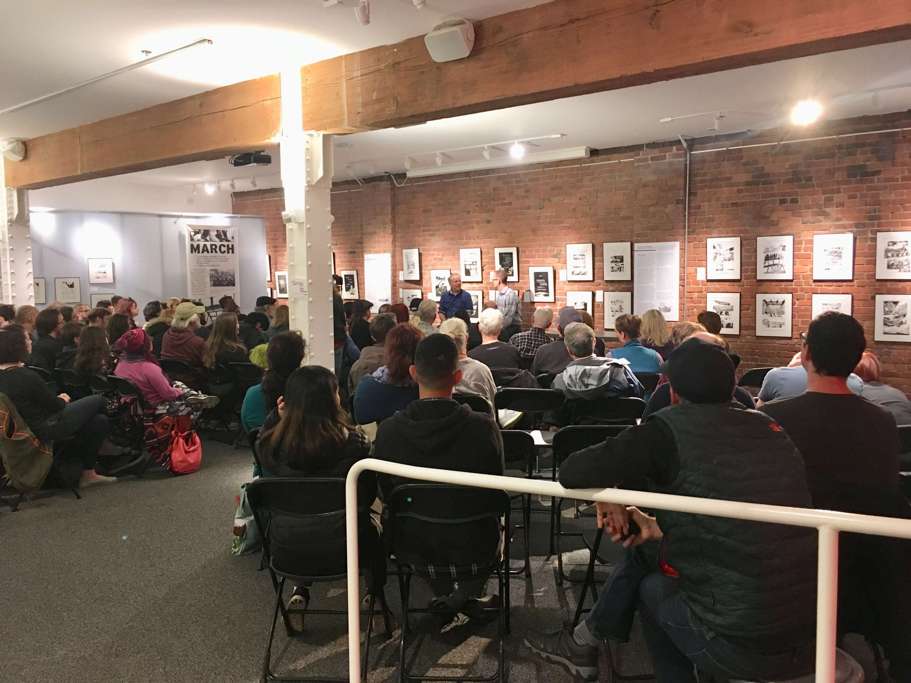 A large audience sits in a gallery space, facing a panel at the front with various framed artworks displayed on the brick walls.