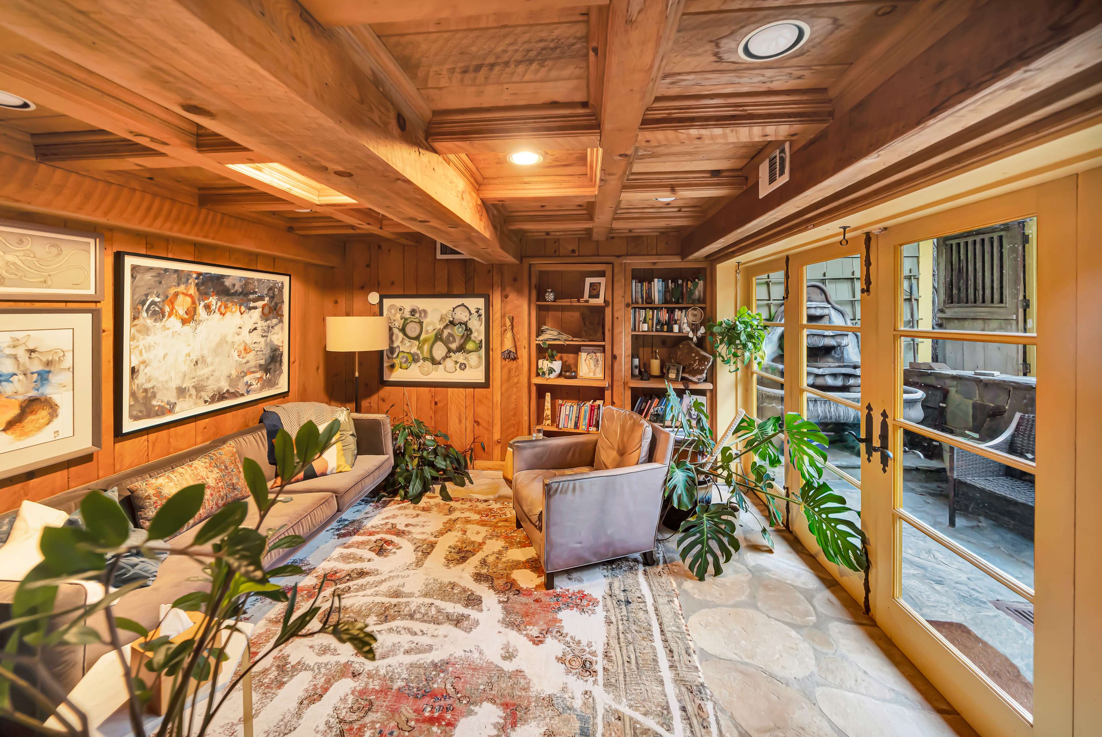 The image shows a cozy room with wooden beams, a sofa, a lamp, bookshelves filled with books, and large glass doors leading to a stone patio with plants.