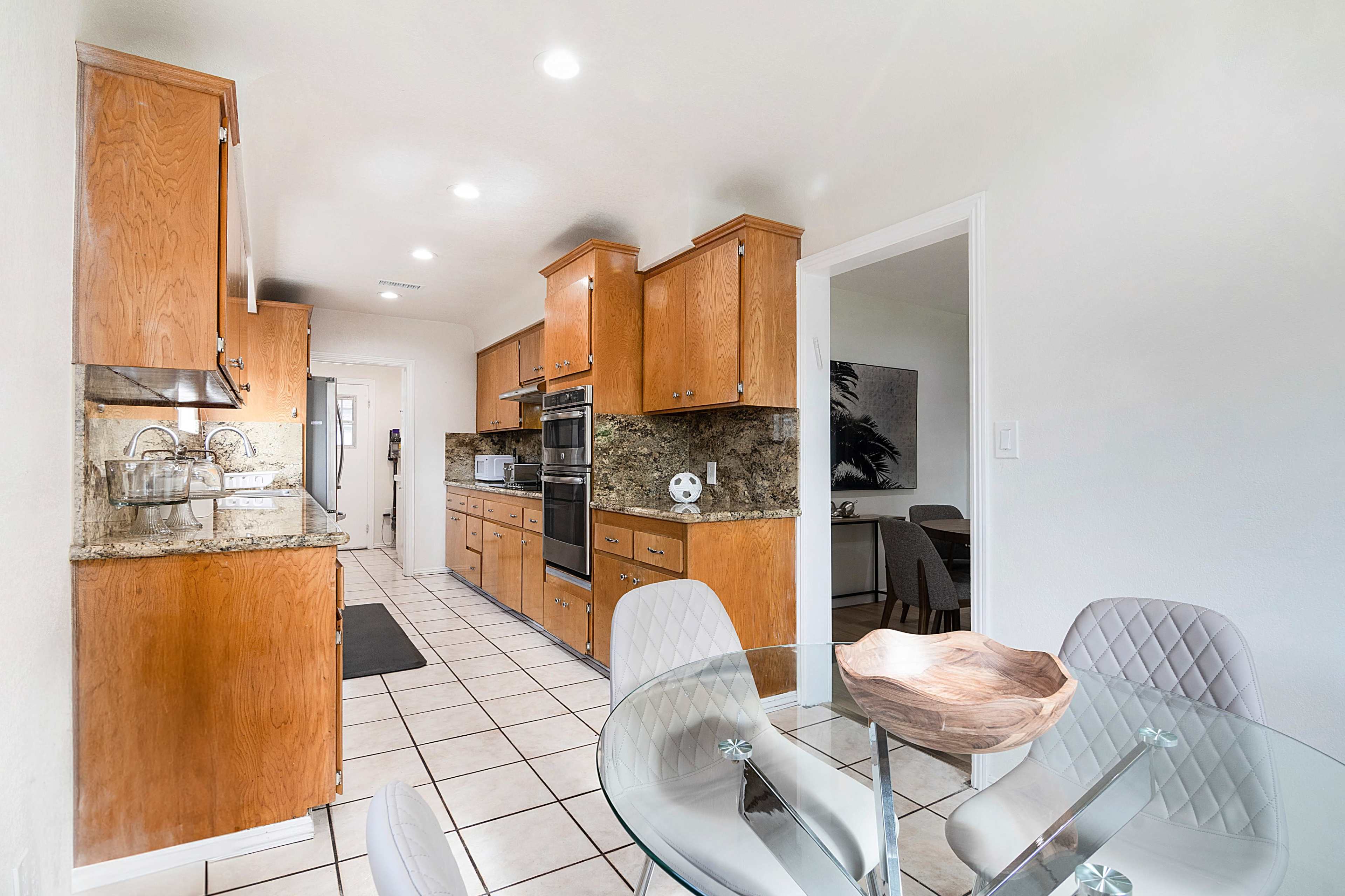 The image shows a modern kitchen featuring wooden cabinets, granite countertops, and a glass dining table with grey chairs.