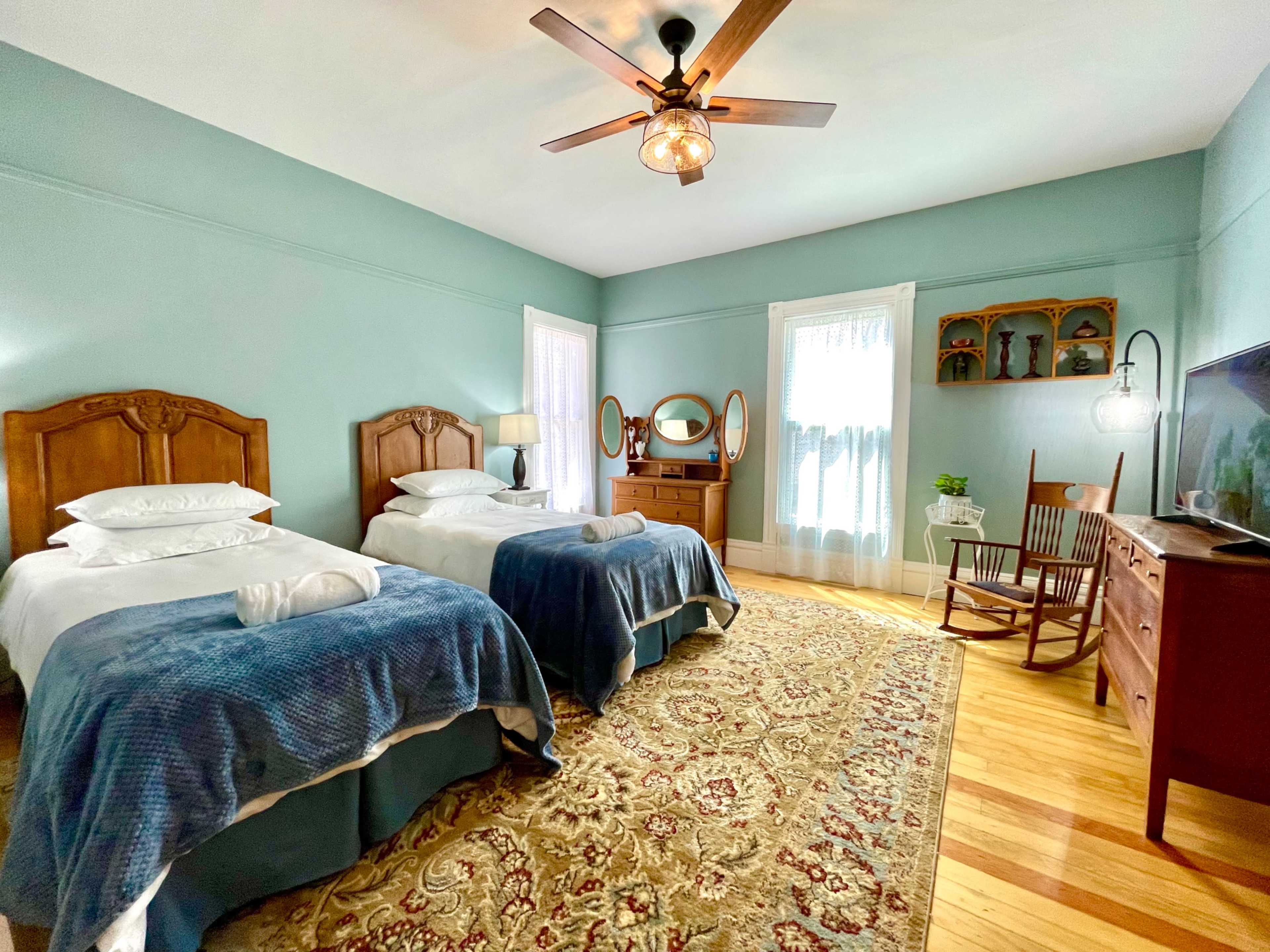A light blue bedroom with two twin beds, a wooden dresser, a rocking chair, and a window with sheer curtains.