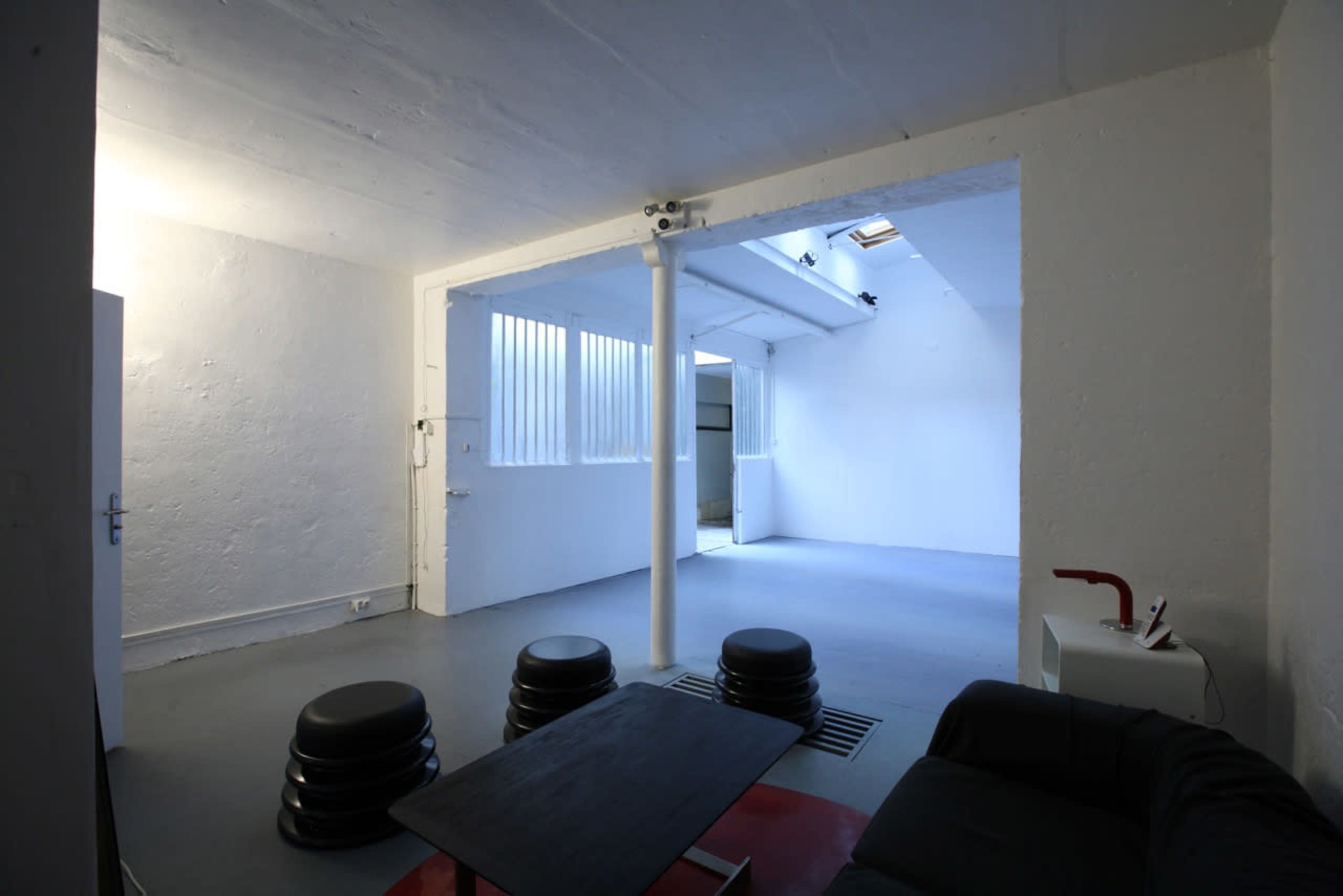 A minimalist, open space with concrete walls, a gray floor, and a few black stools arranged around a low table.