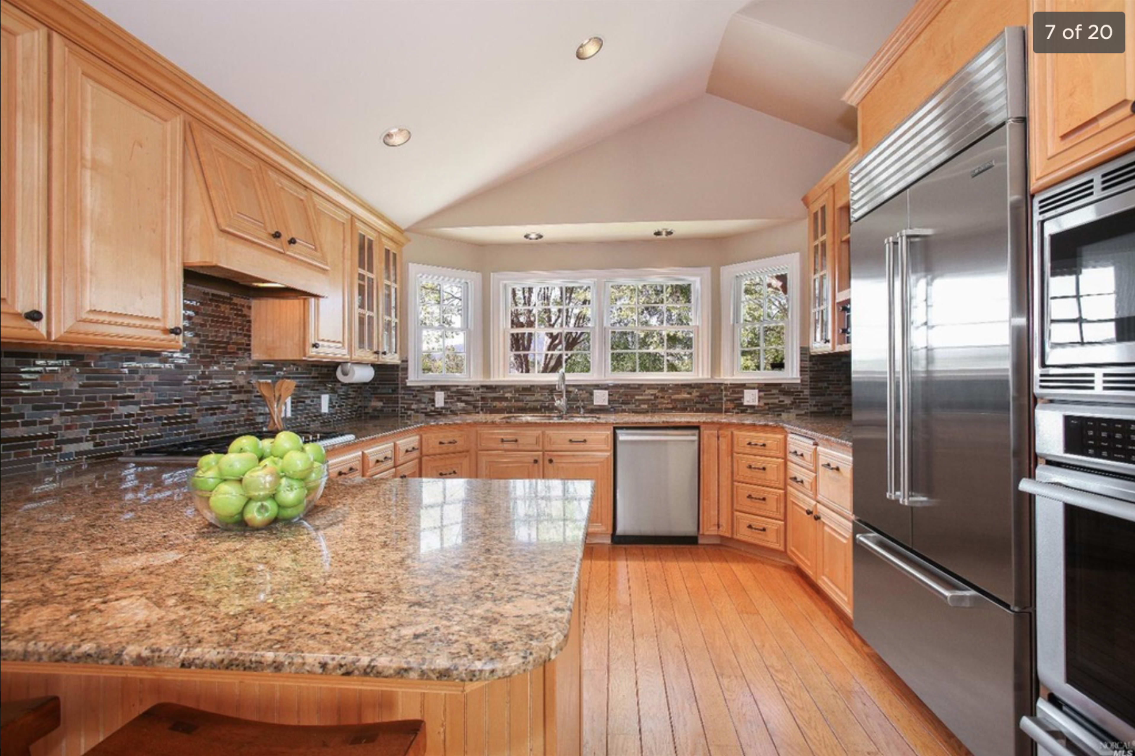 A kitchen features wooden cabinets, a granite countertop with a bowl of green apples, and stainless steel appliances under a sloped ceiling.