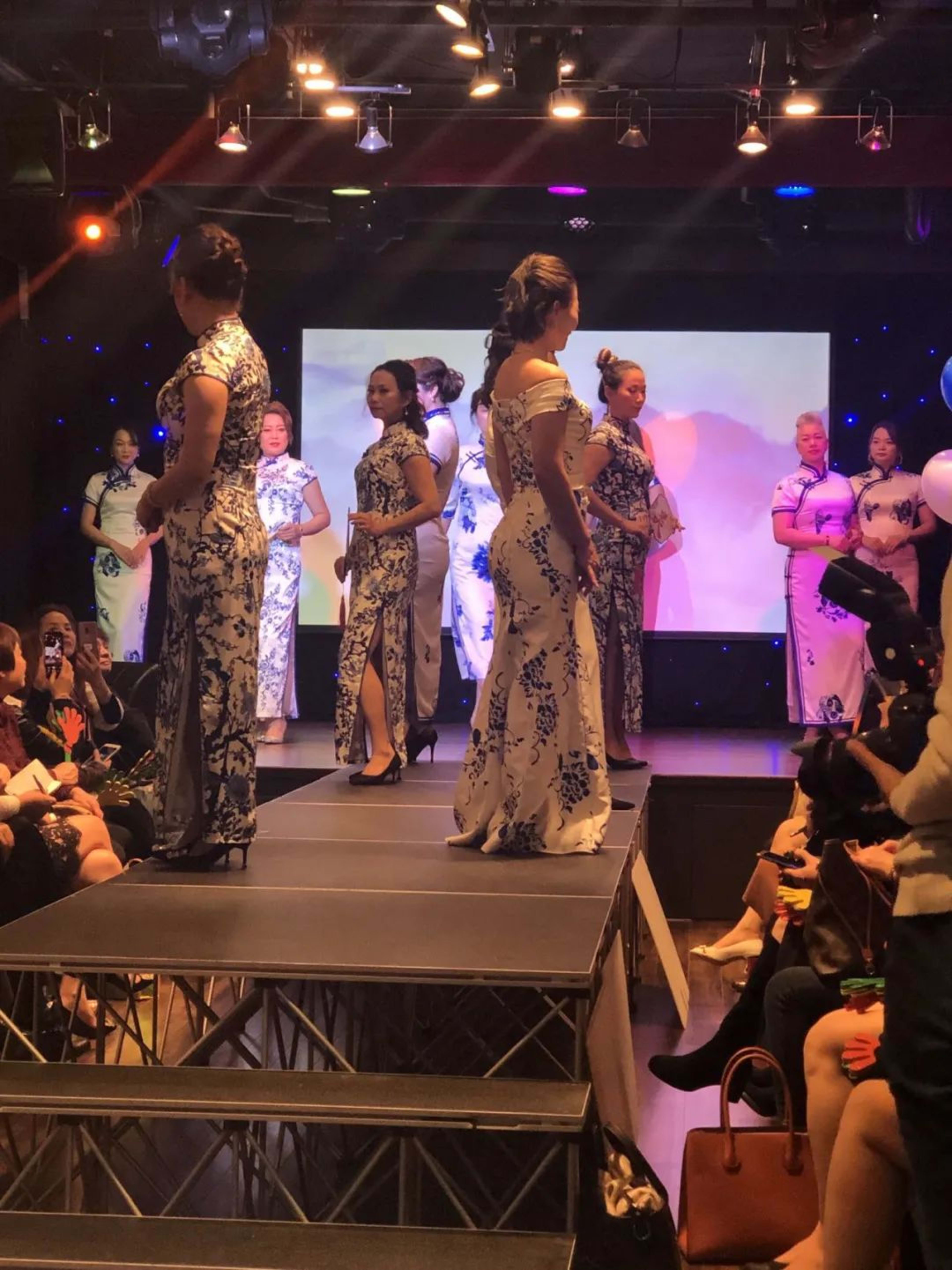 Models wearing traditional dresses walk down a runway while an audience observes.