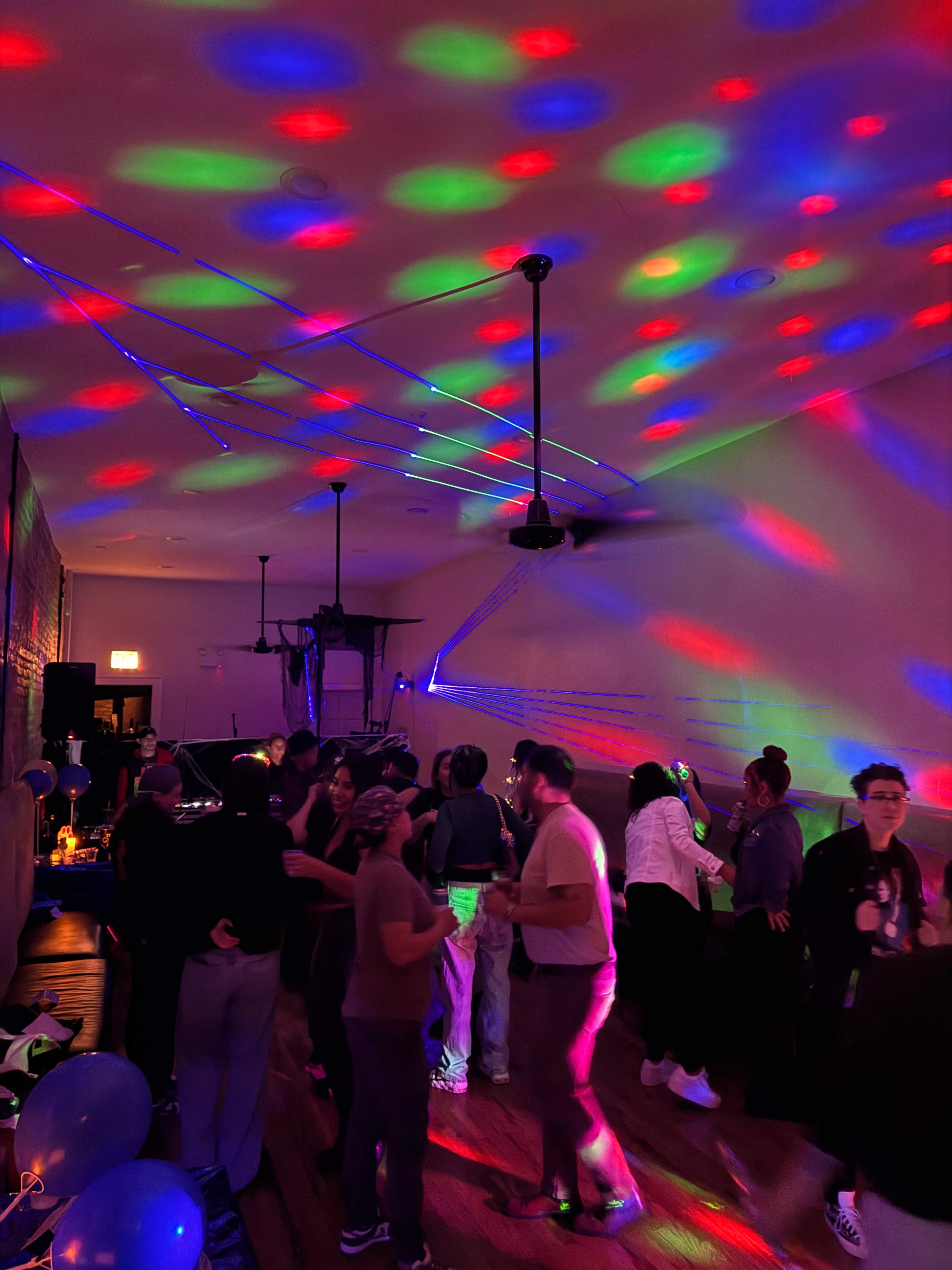 A group of people is dancing in a dimly lit room illuminated by colorful lights and laser effects.