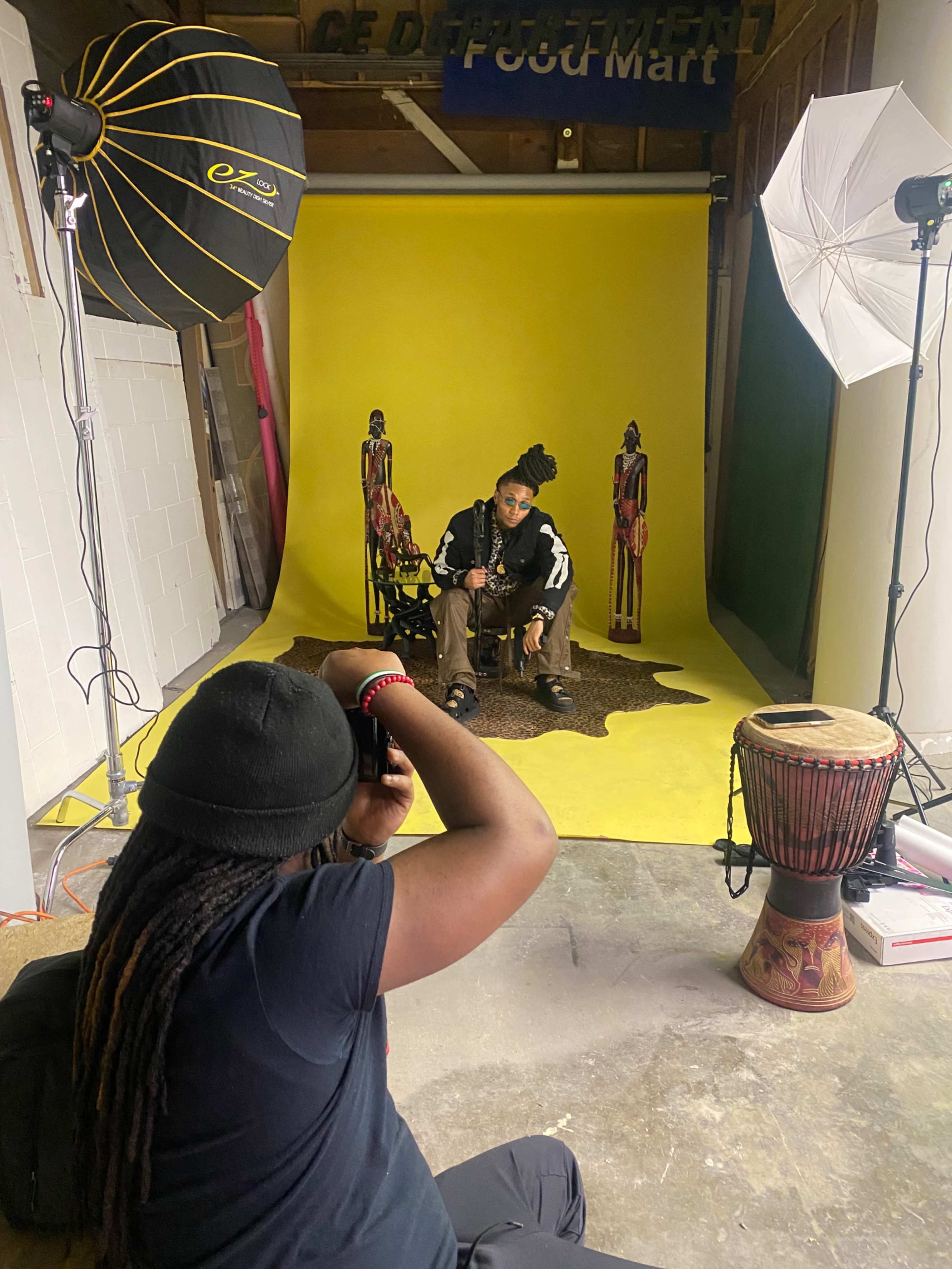 A photographer captures a model posing on a yellow backdrop surrounded by decorative figures and a drum in a studio setting.
