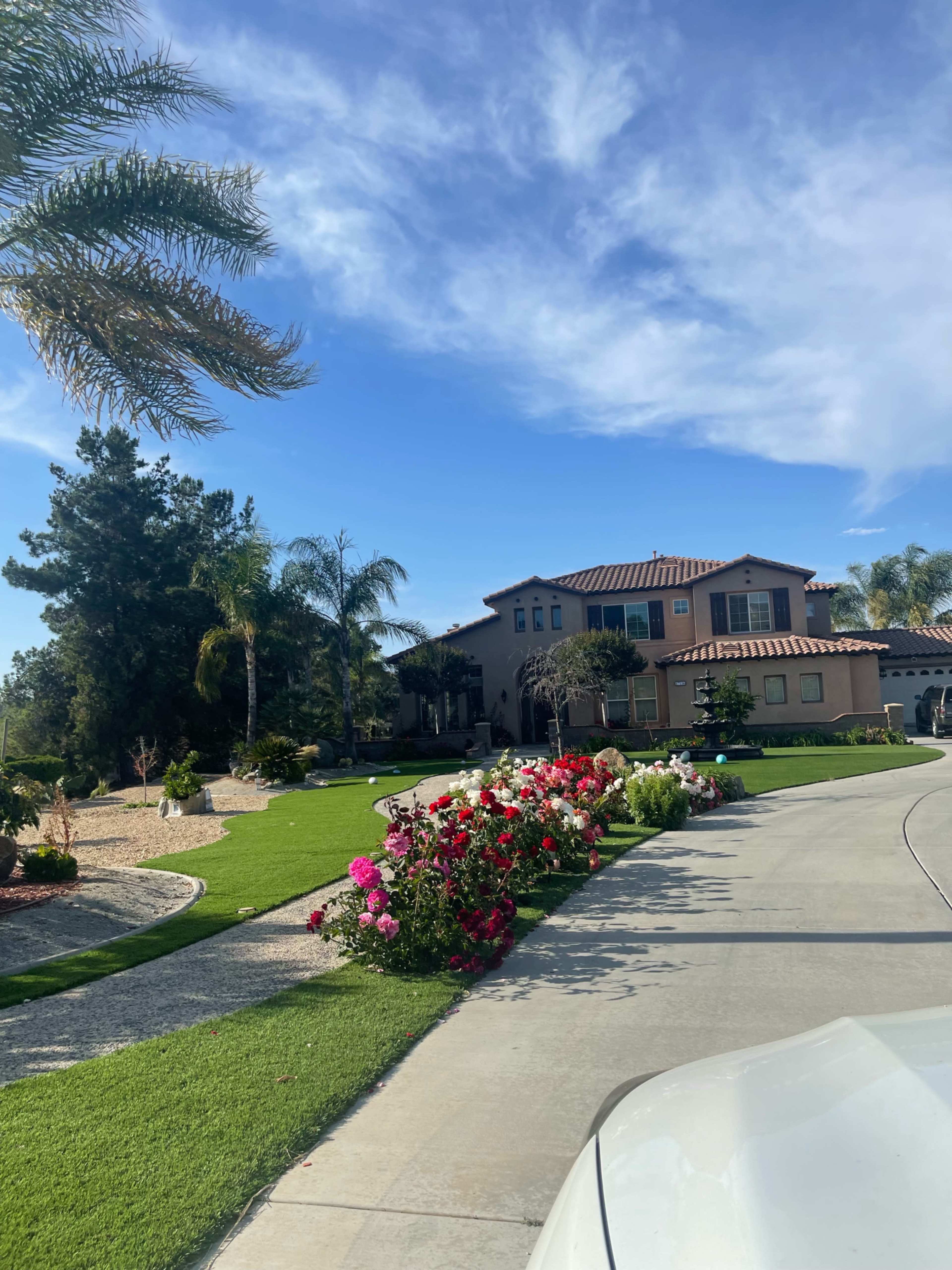 A large house with a tiled roof is set back from a curved driveway lined with vibrant flower beds and palm trees under a blue sky.
