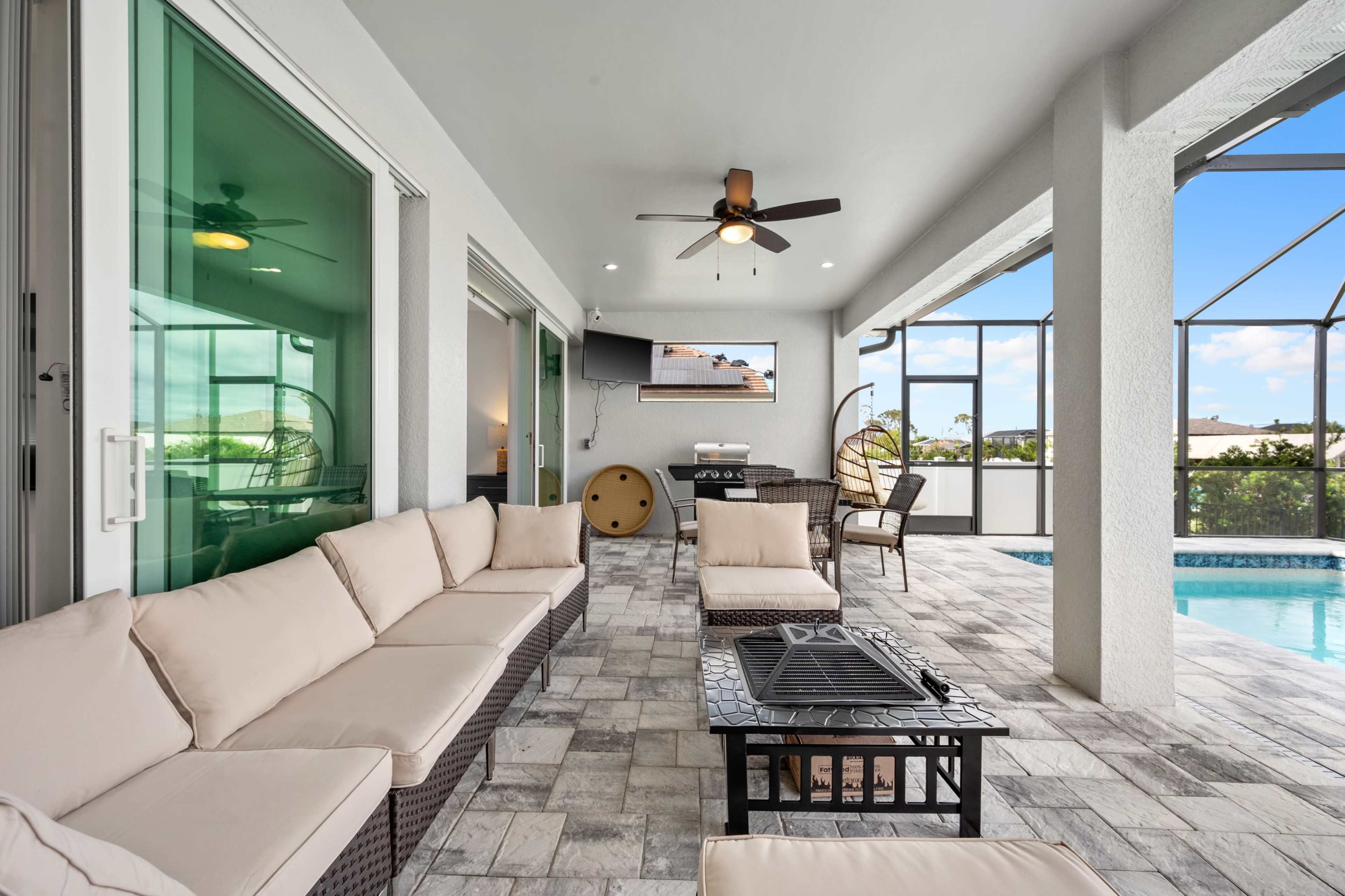 A modern outdoor patio with a sectional seating arrangement, a fire pit, and a television mounted on the wall, overlooking a pool area enclosed by a screen.