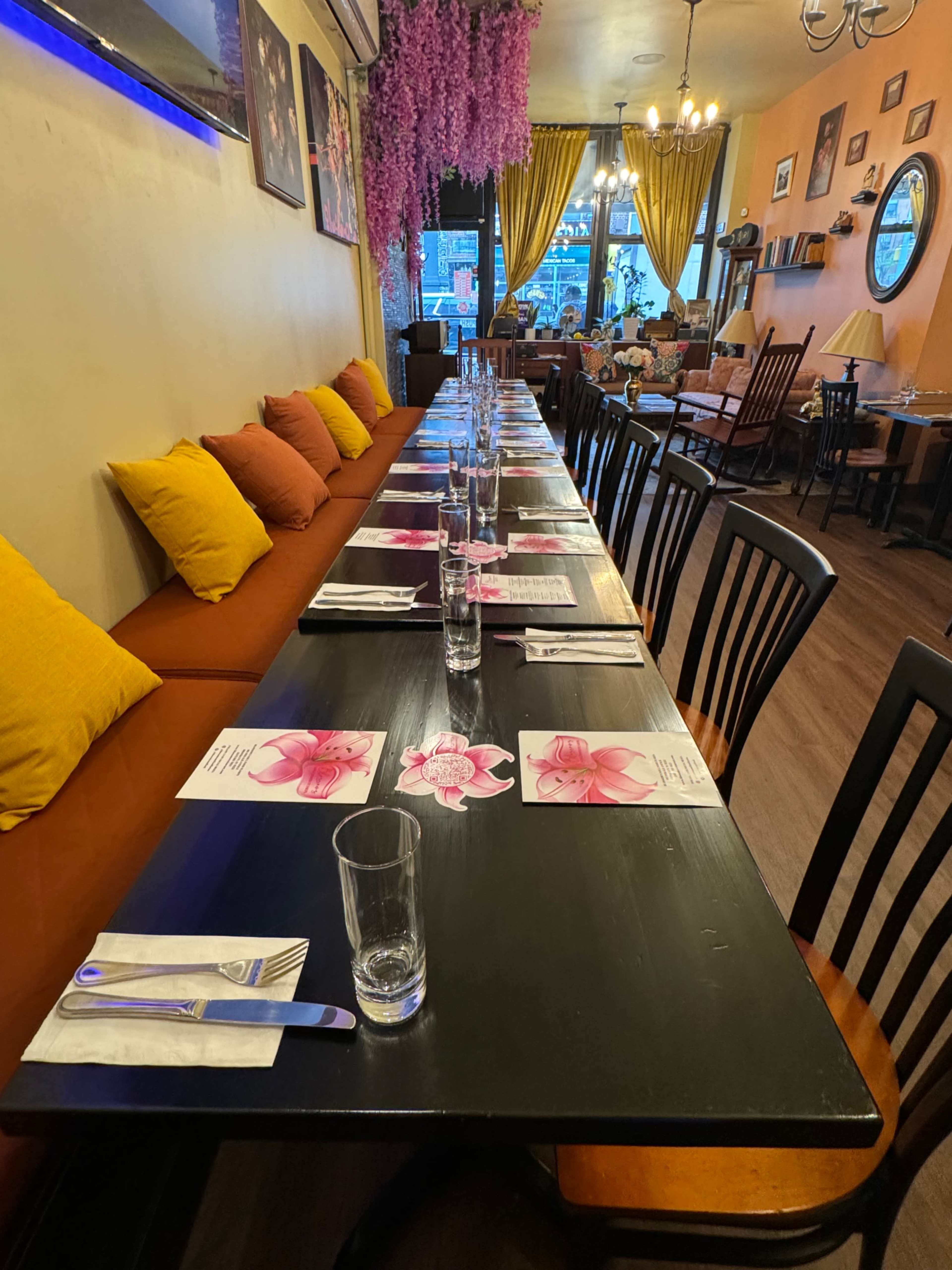 A long dining table is set with water glasses and floral menus, surrounded by colorful cushions in a cozy restaurant.
