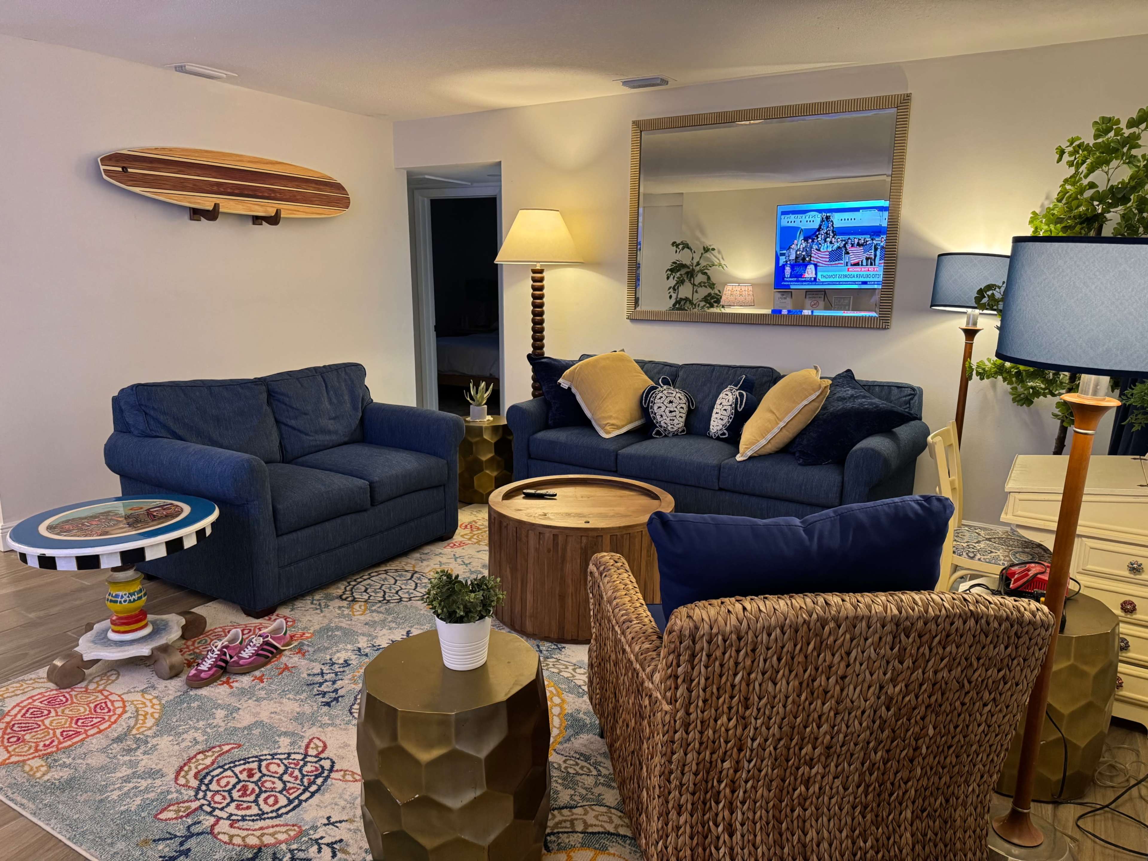 The scene depicts a cozy living room featuring two blue sofas, a round wooden coffee table, a decorative area rug, and a large mirror, with a television visible in the background.