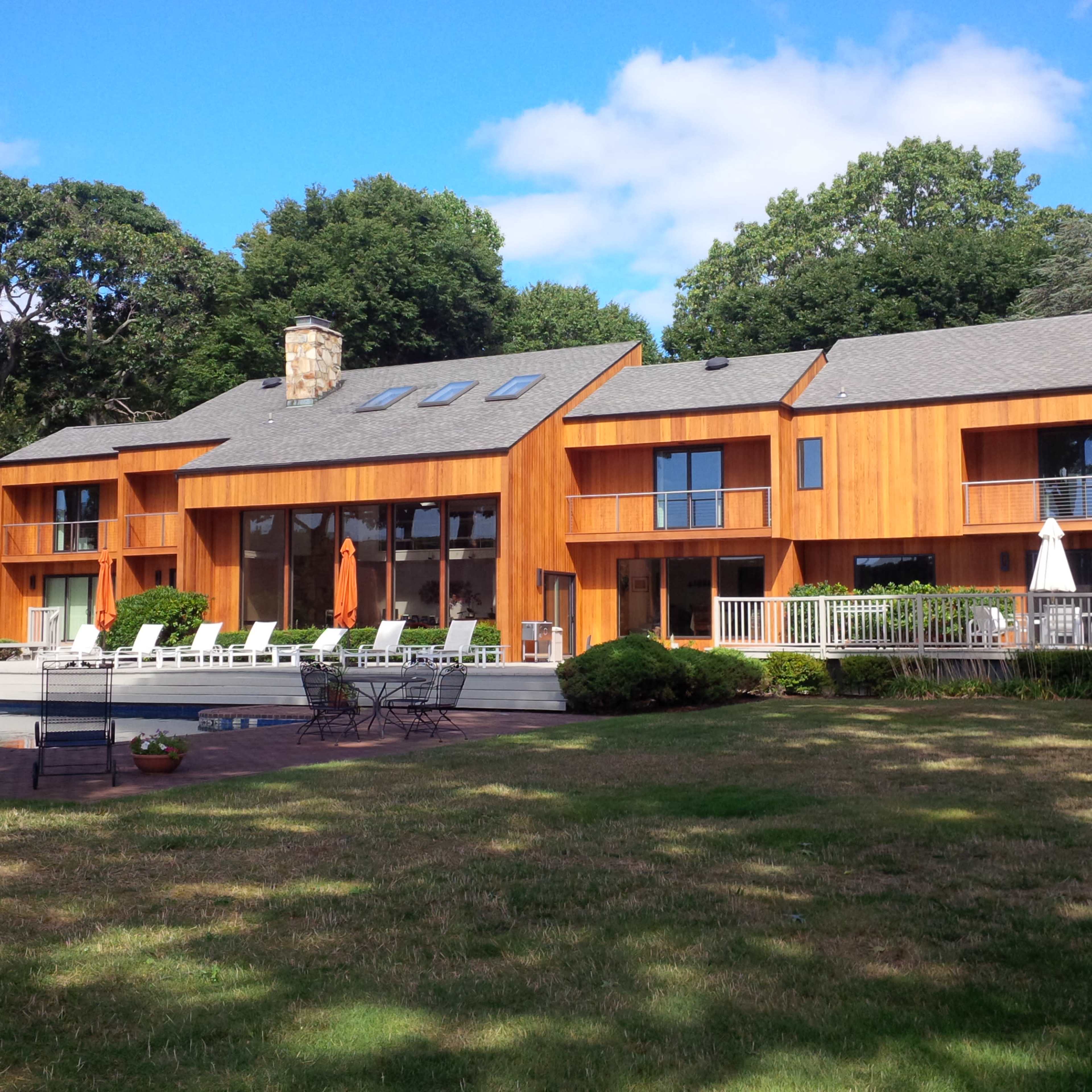 A modern wooden house with large windows and a deck is surrounded by a green lawn and trees.