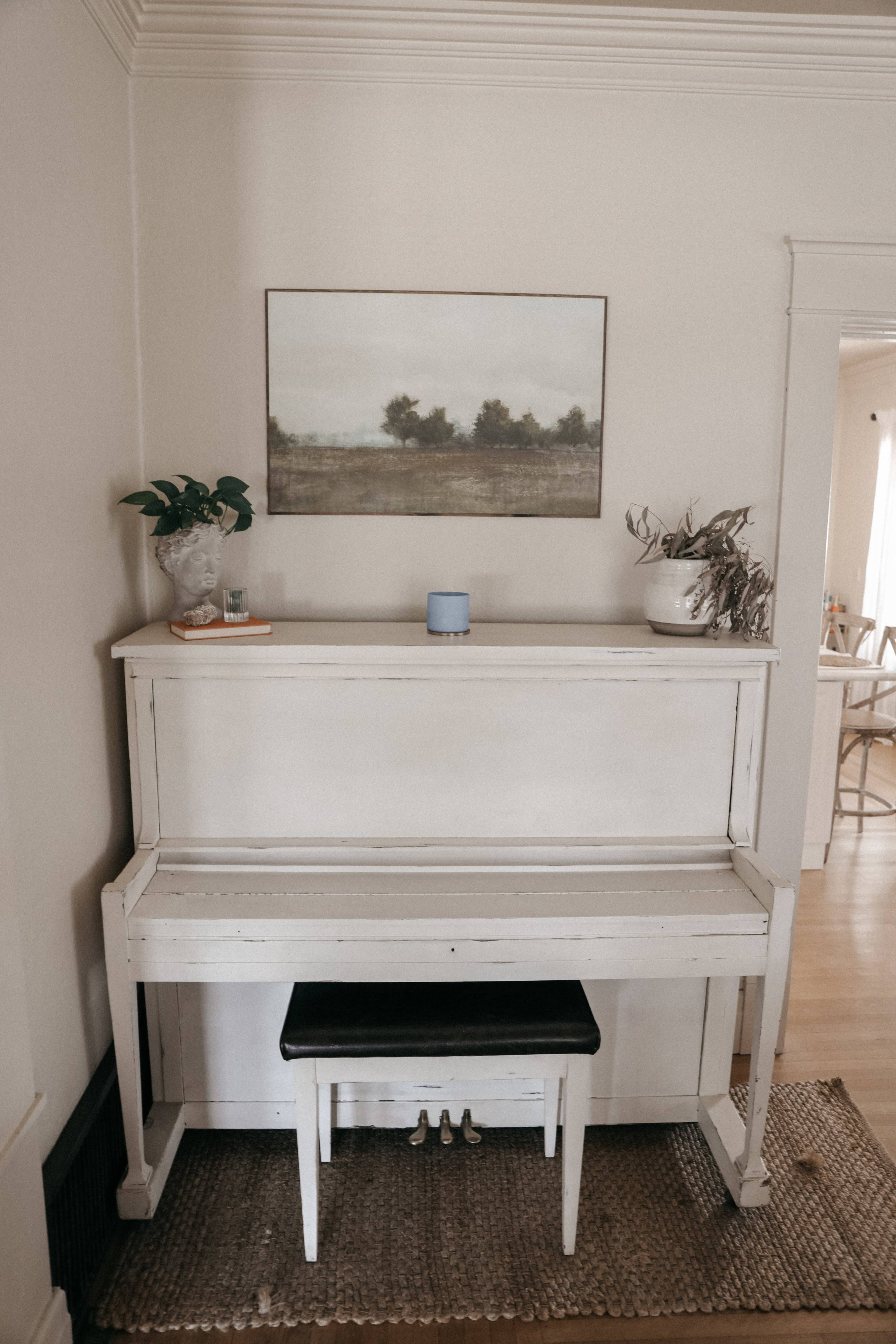 A white piano with a black bench is positioned against a light-colored wall, adorned with a painting and potted plants.