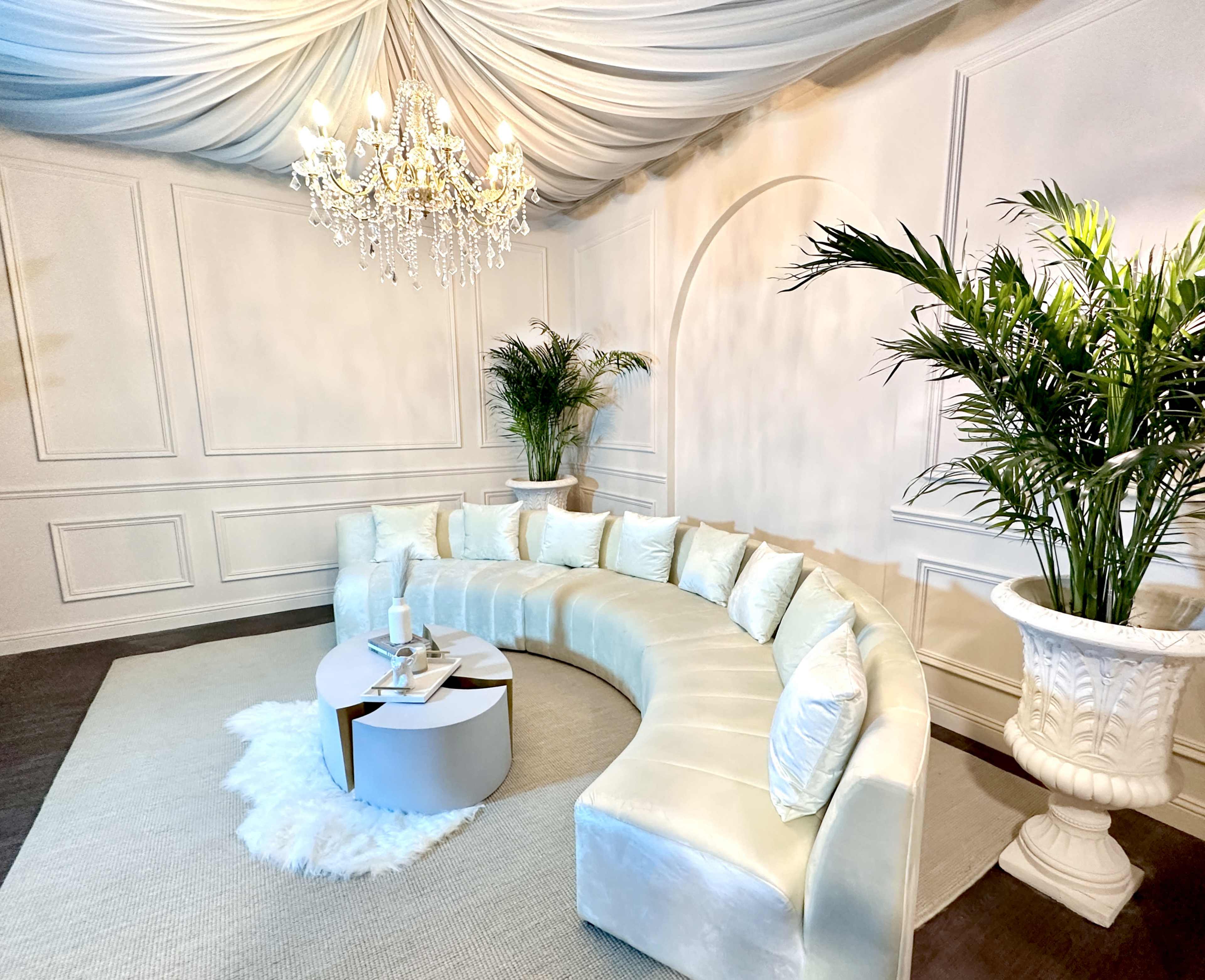 The image shows a spacious, elegantly furnished room with a curved white couch, a round table, a chandelier, and decorative plants.