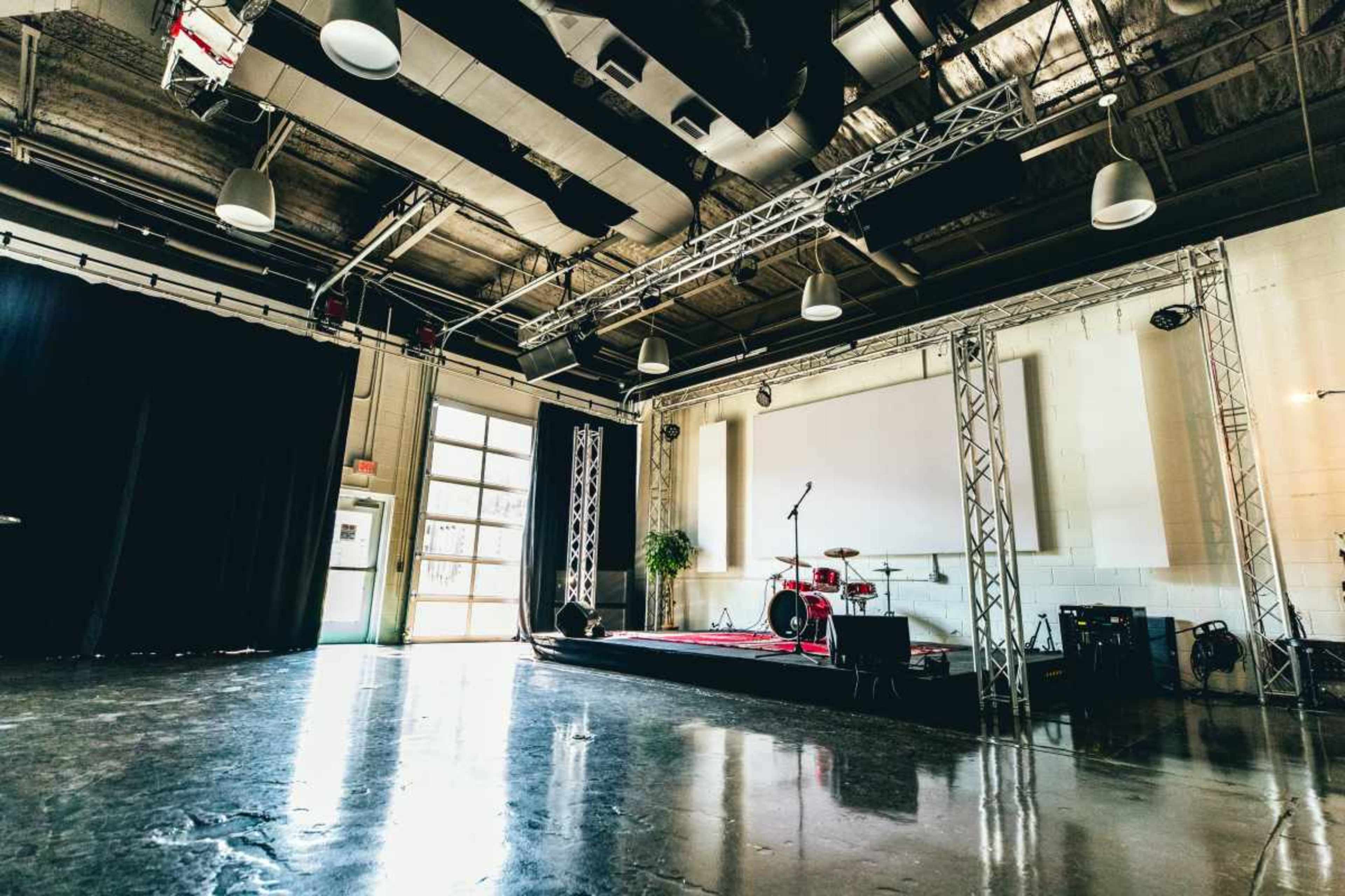 The image shows an empty rehearsal space with a small stage set up for a band, featuring a drum kit and lighting fixtures, along with a large open door leading outside.