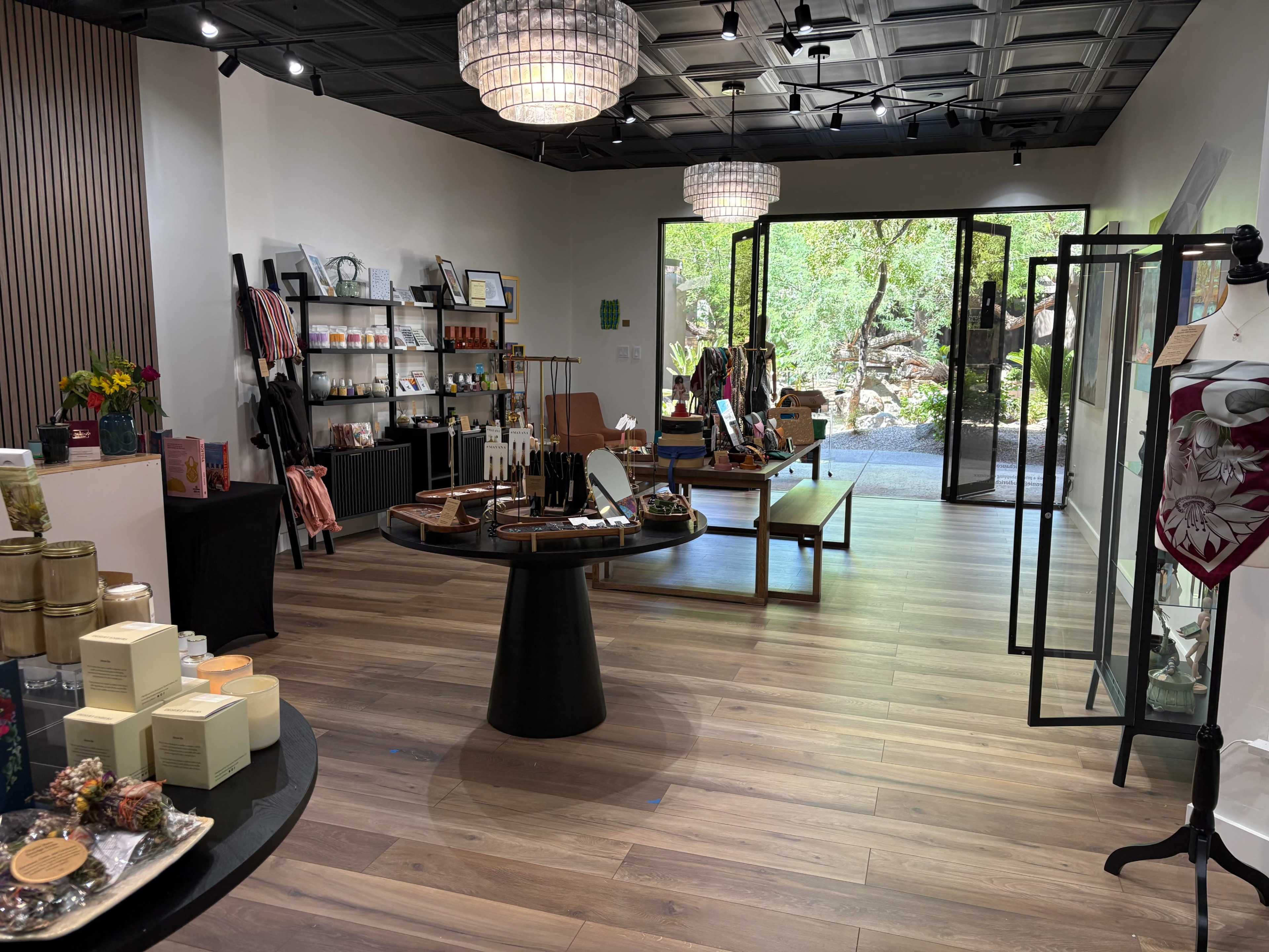 The image shows a well-organized retail store featuring a variety of products displayed on tables and shelves, with large windows revealing a lush green outdoor area.