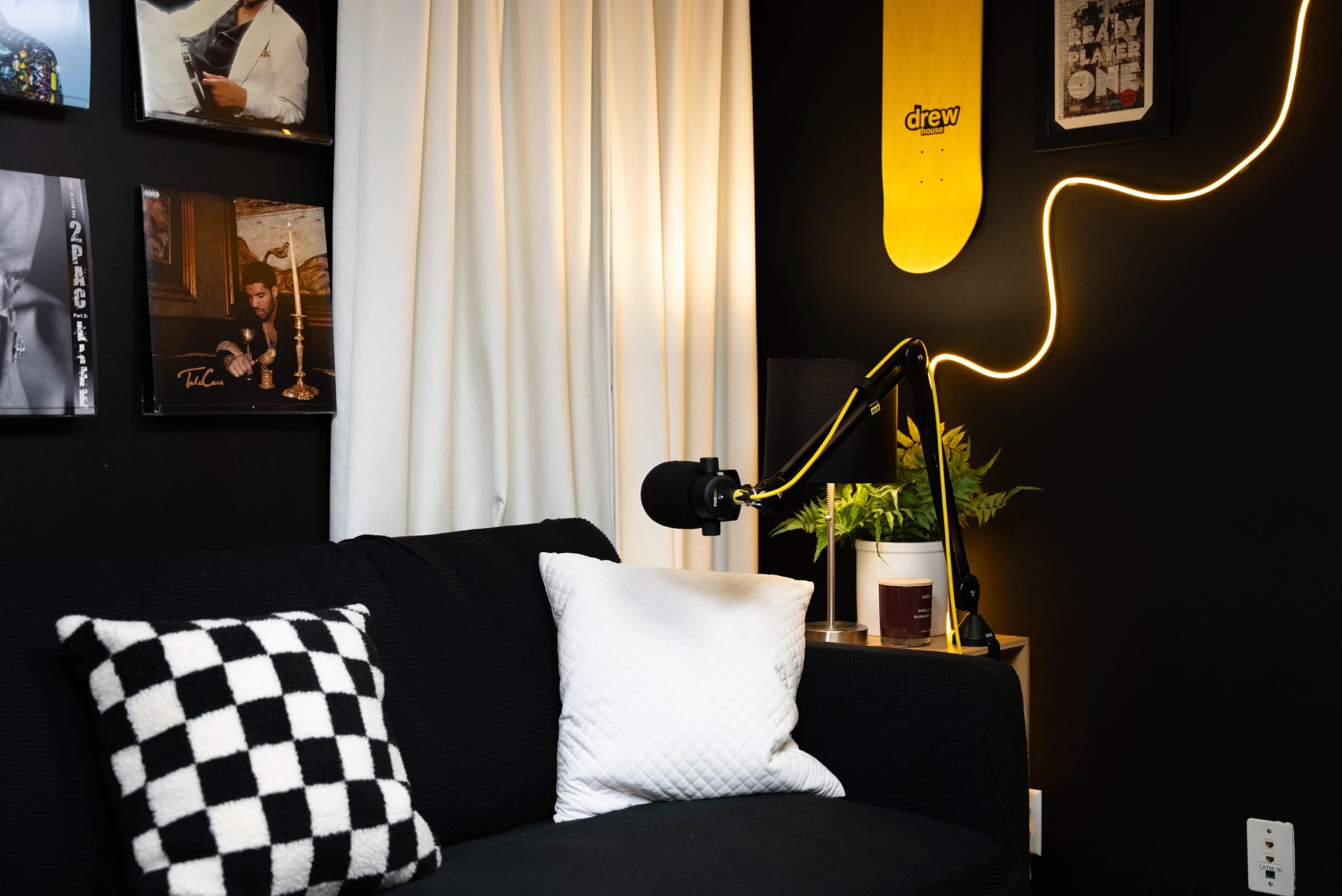 A black sofa with checkered and white pillows is positioned near a wall displaying framed artwork and a skateboard, illuminated by a light fixture and a microphone stand.