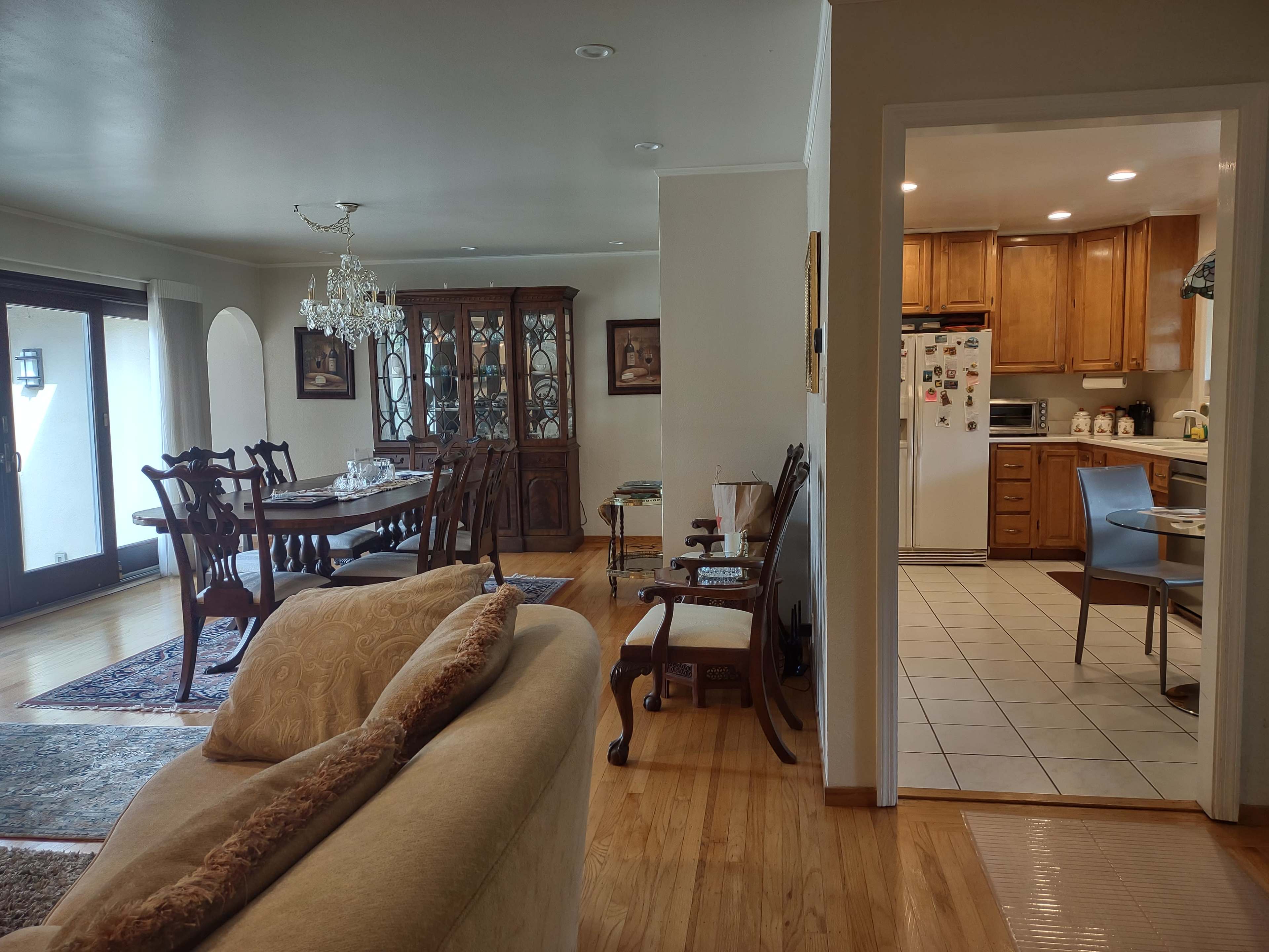 A spacious dining and living area with a wooden dining table and a kitchen visible through an open doorway.