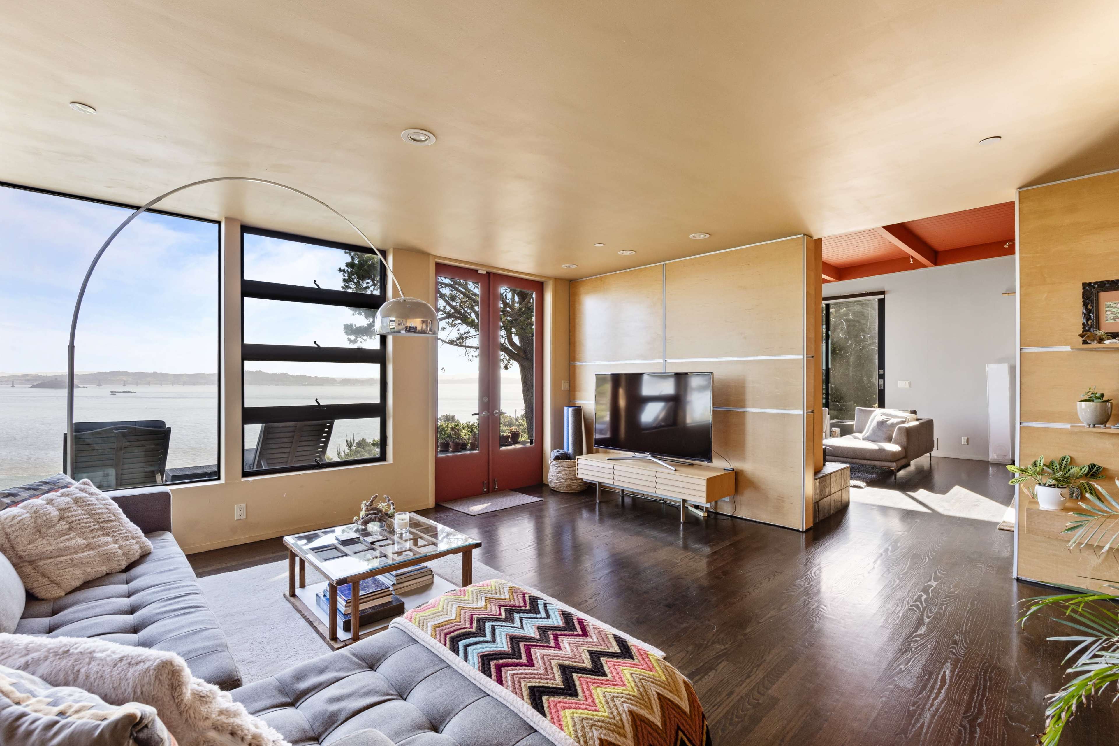 A modern living room features a sofa, a television mounted on a wooden media console, and large windows showcasing a waterfront view.