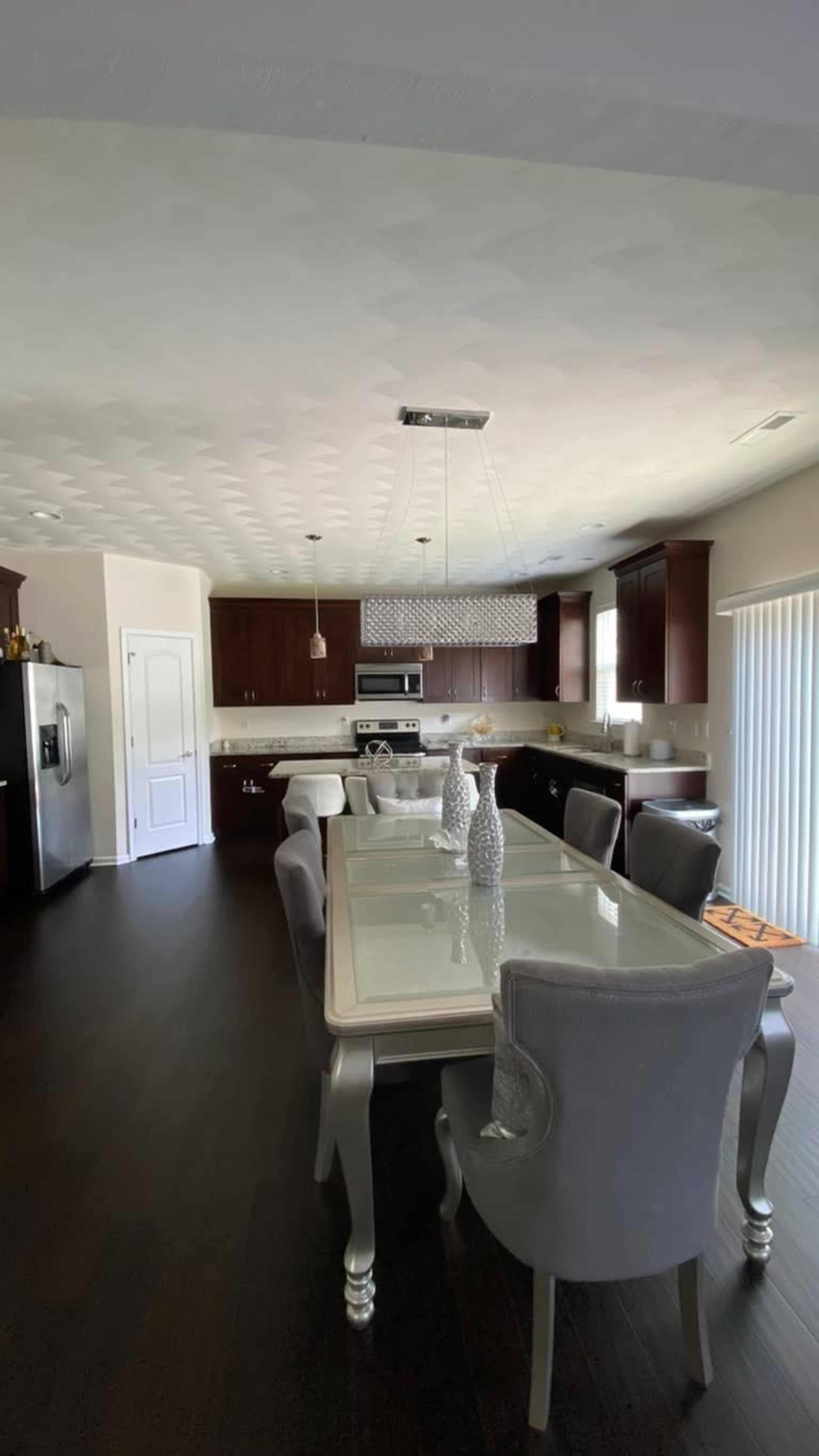 A modern kitchen and dining area featuring a rectangular glass dining table surrounded by upholstered chairs, with dark wood cabinetry and stainless steel appliances.