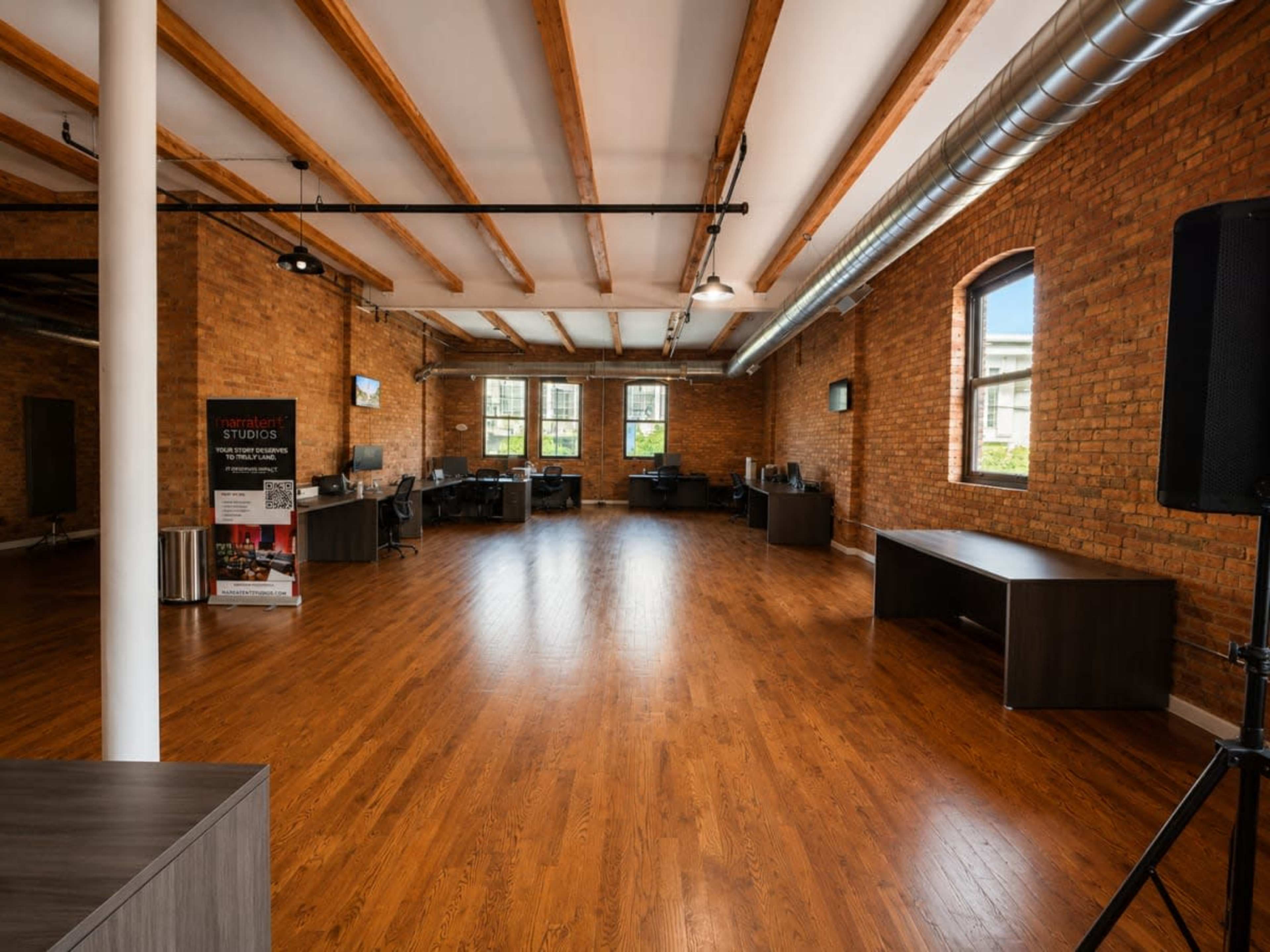 The image shows a spacious, modern studio with wooden floors, exposed brick walls, and several workstations equipped with computers.