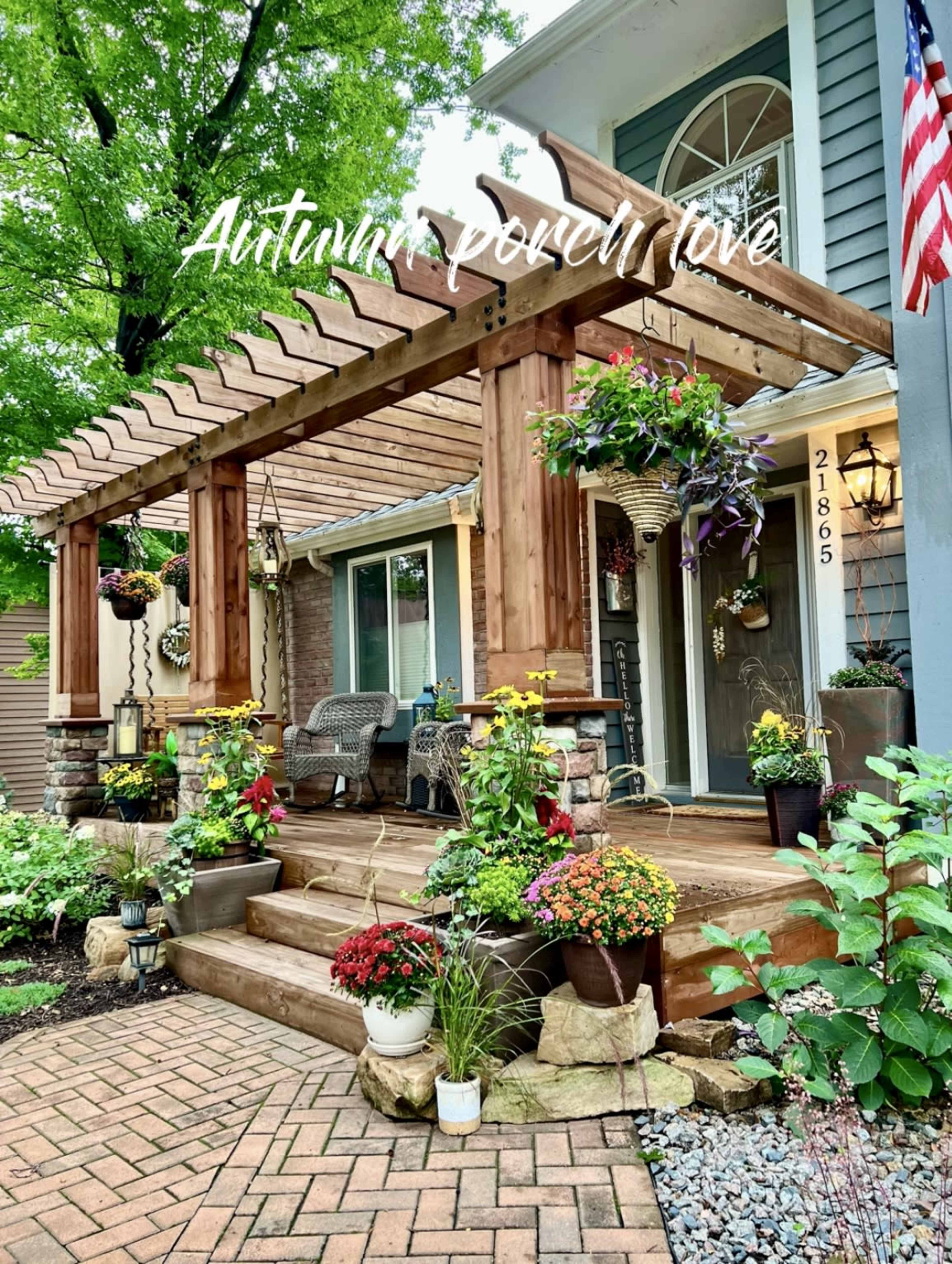 A front porch with a wooden pergola adorned with hanging flower pots and a variety of potted plants arranged on the steps and walkway.