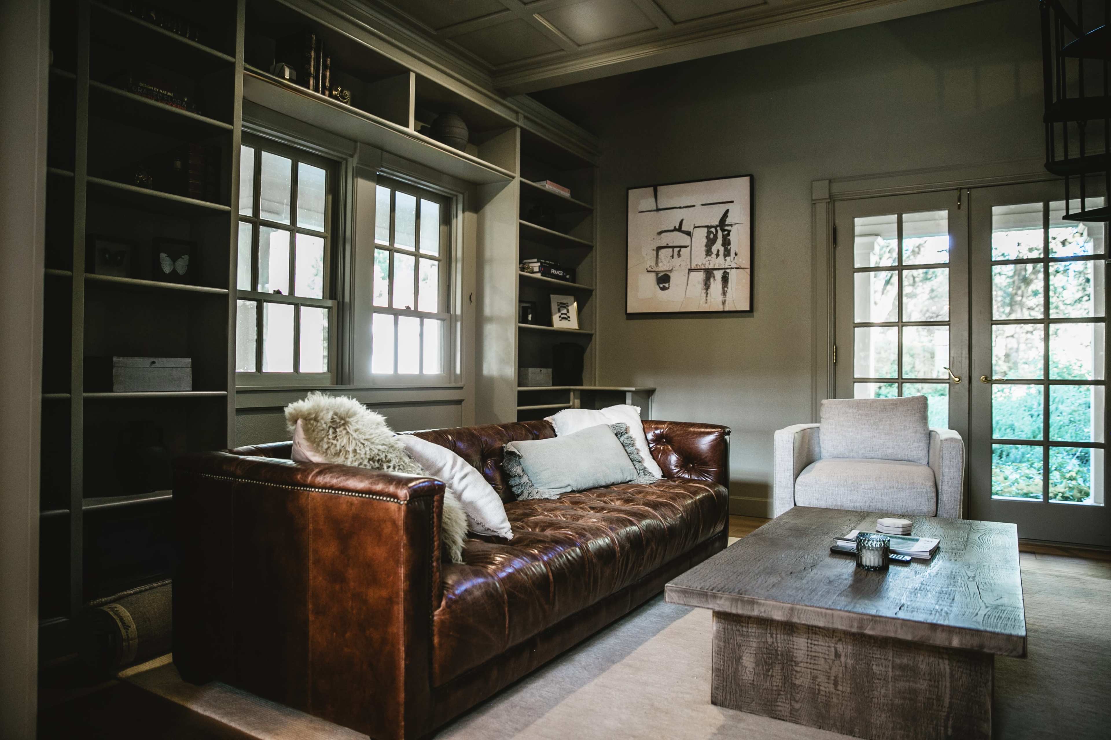 The image shows a cozy living room featuring a brown leather sofa, a light gray armchair, and a wooden coffee table, all arranged around a large window that opens up to an outdoor view.