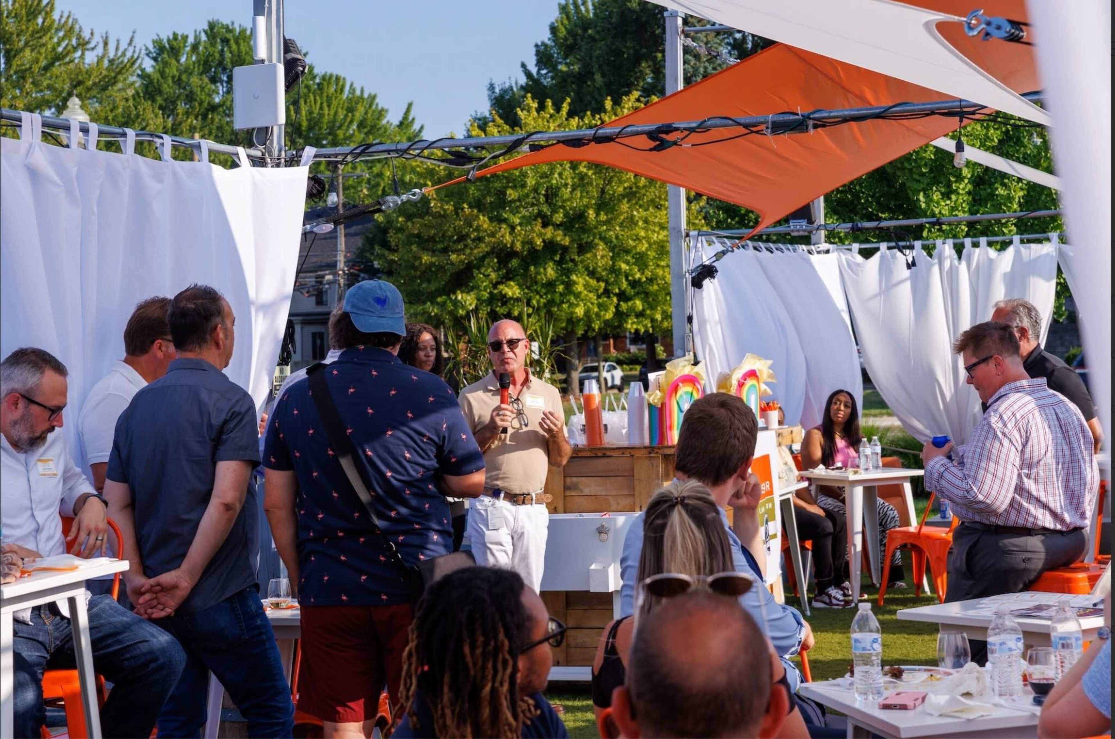A group of people is gathered outdoors in a tented area, with one man speaking to the crowd while others listen attentively.