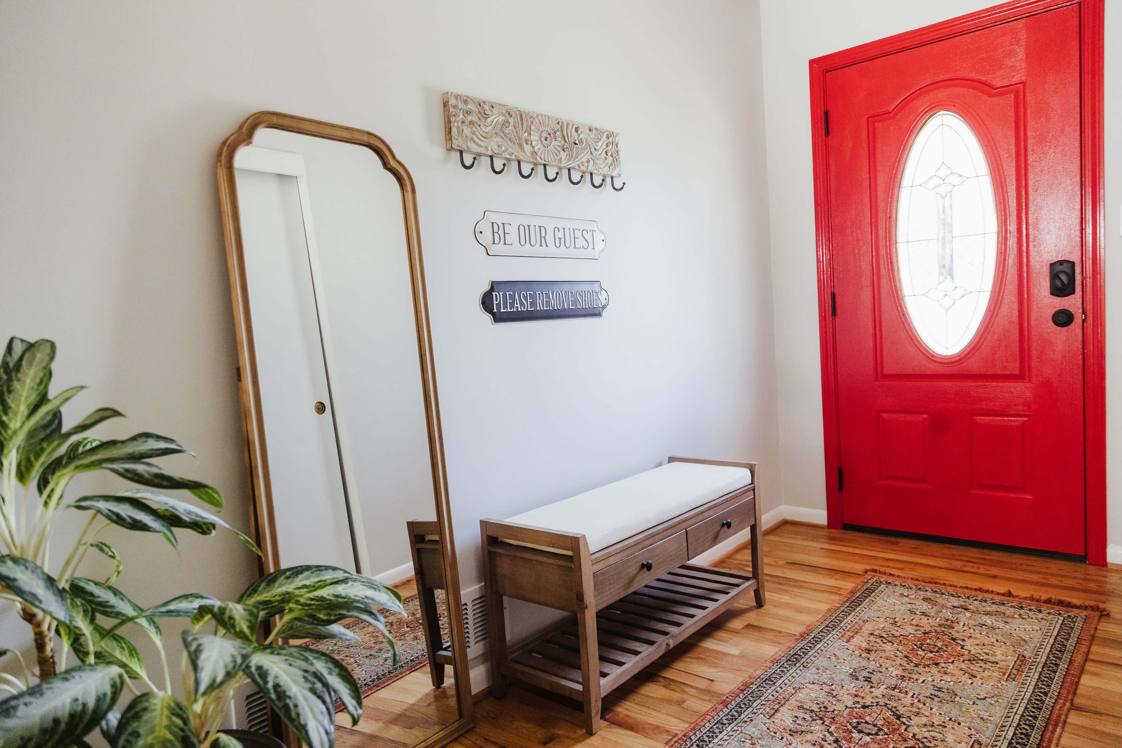 An entryway with a red front door, a wood bench with a cushion, a tall mirror, a decorative wall sign, and a patterned rug.
