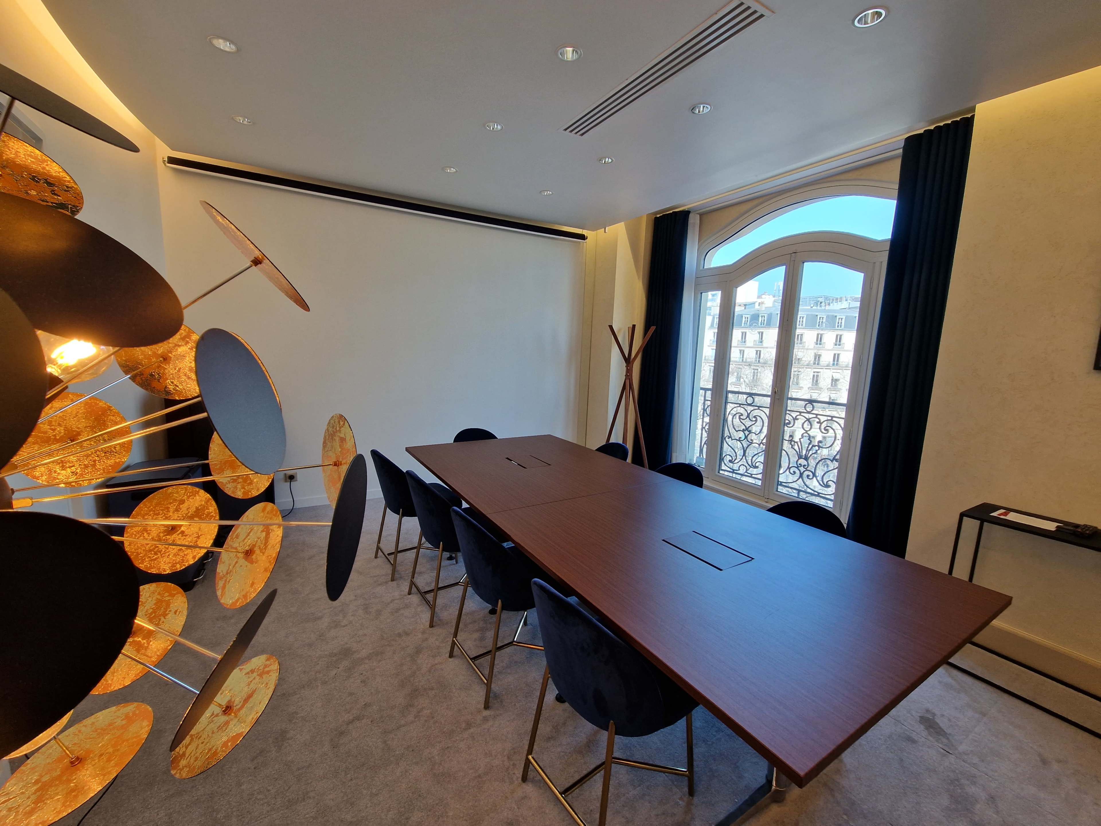 A modern conference room with a large table surrounded by black chairs and a decorative wall feature next to a window overlooking a balcony.