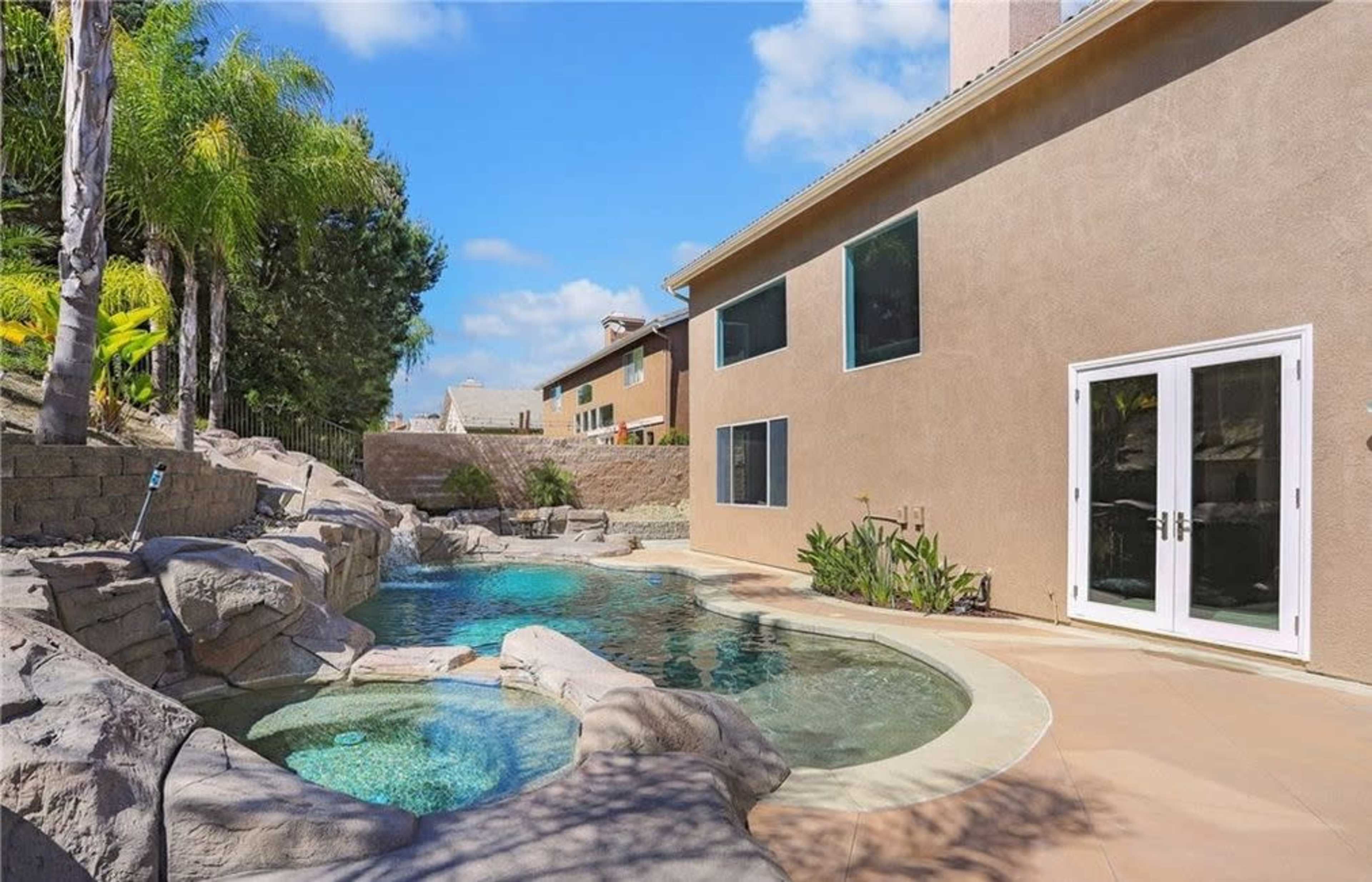 A backyard with a pool and spa area surrounded by rocks and plants, adjacent to a beige house with large windows.