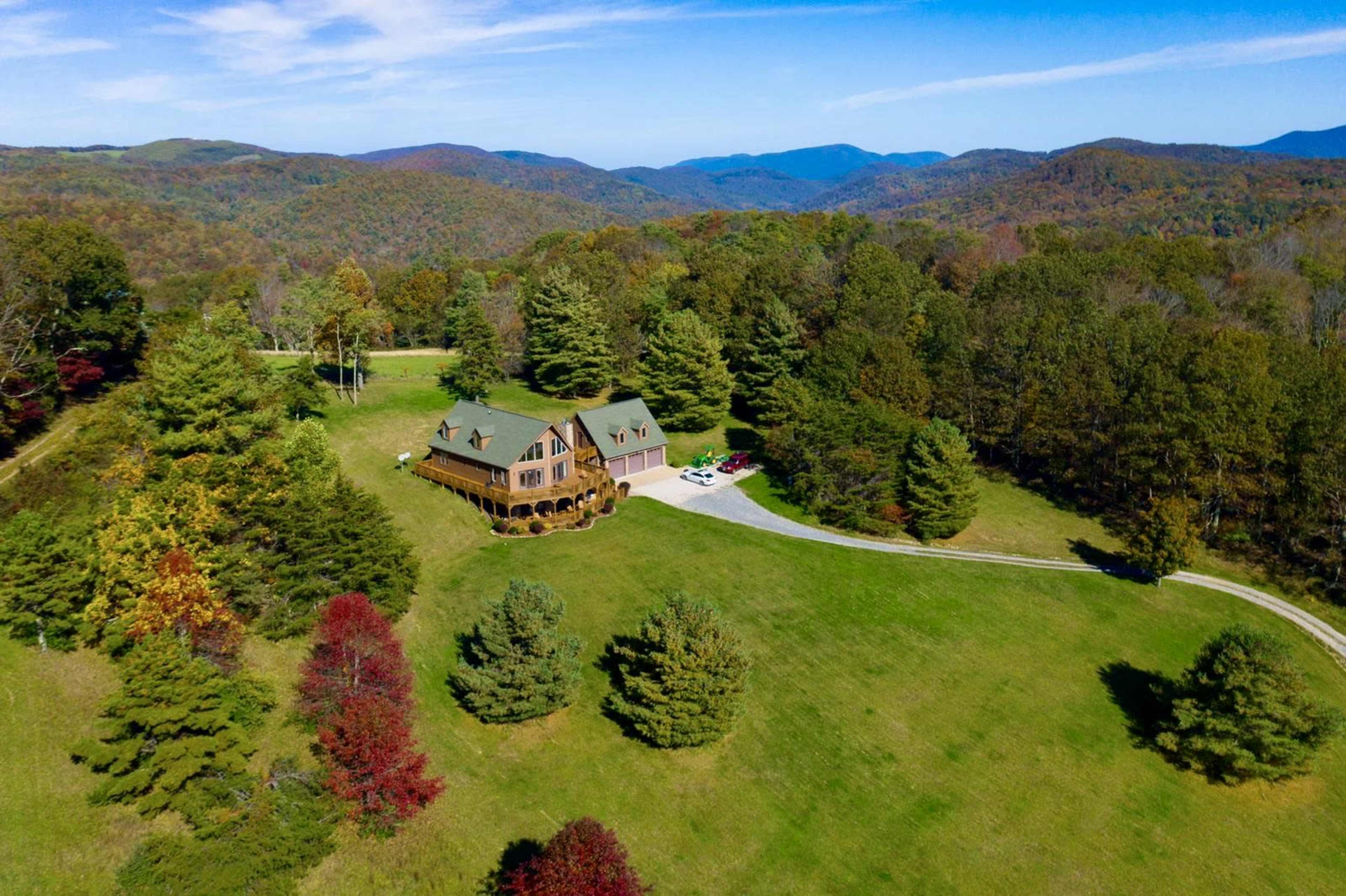 A large, multi-story house is surrounded by rolling hills and trees, with a winding driveway leading to it.