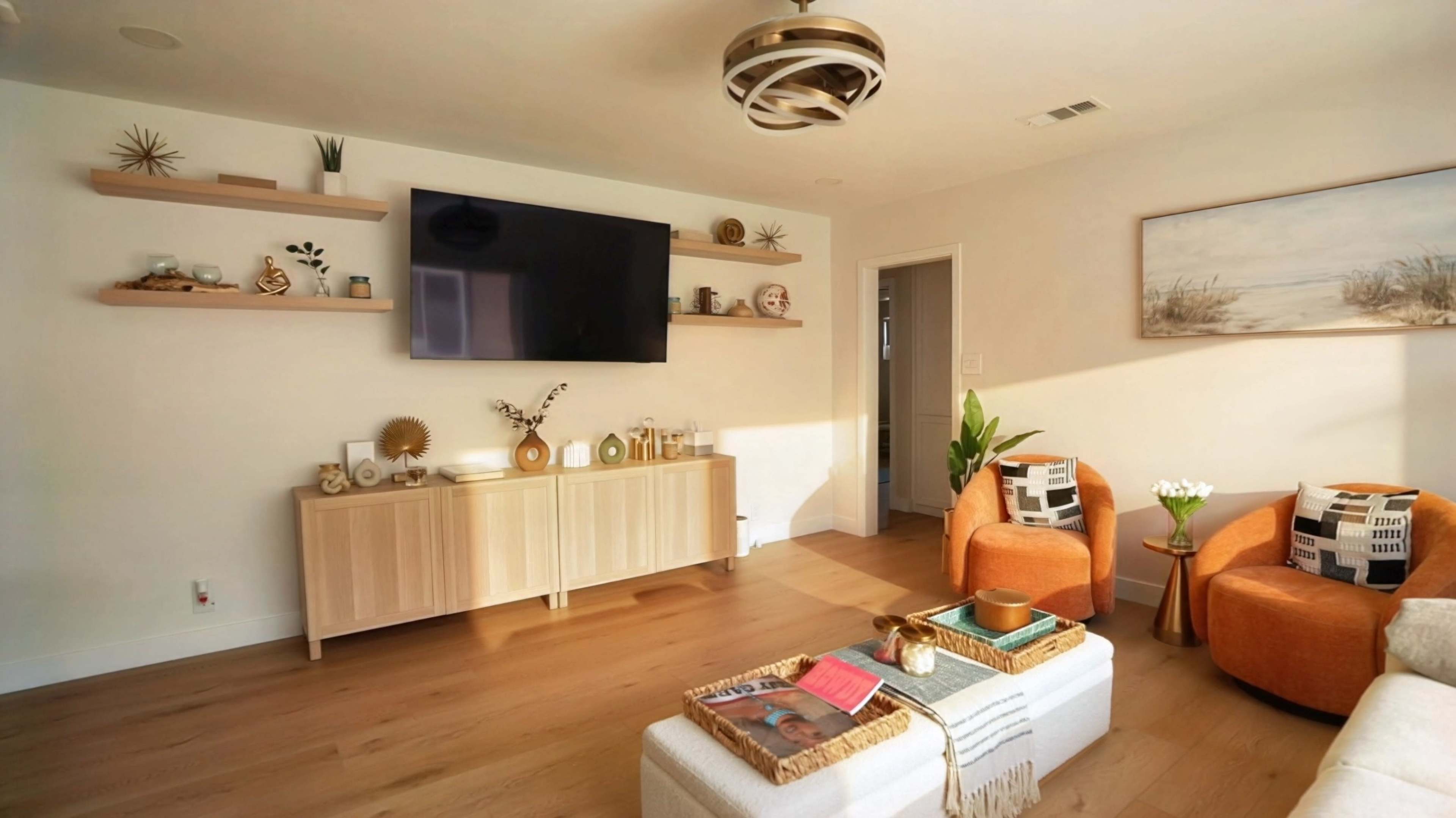 A modern living room features a mounted TV above a light wooden sideboard, complemented by orange armchairs and a coffee table with decorative items.
