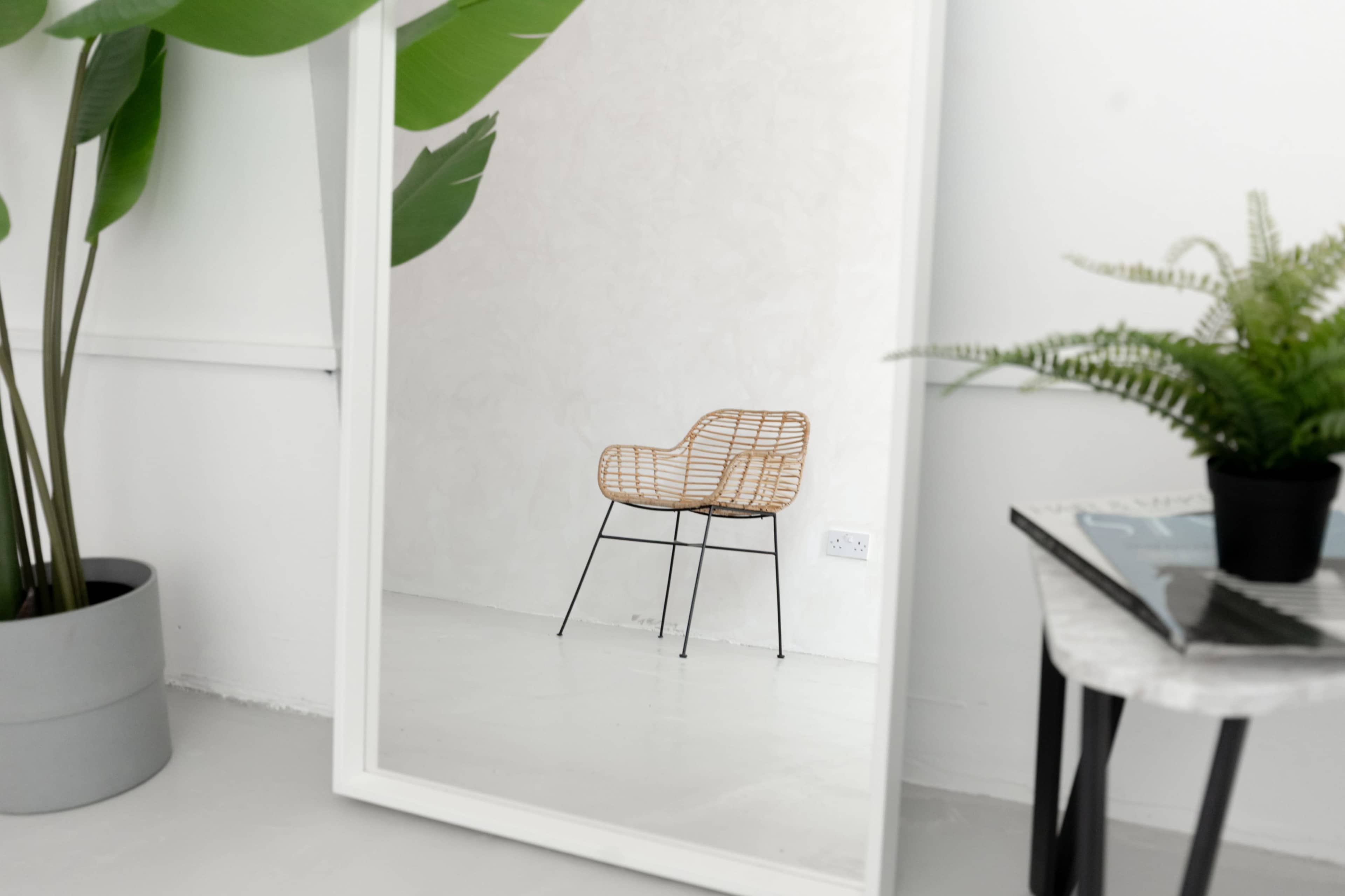 A large mirror reflects a wicker chair and green plants in a minimalist room.