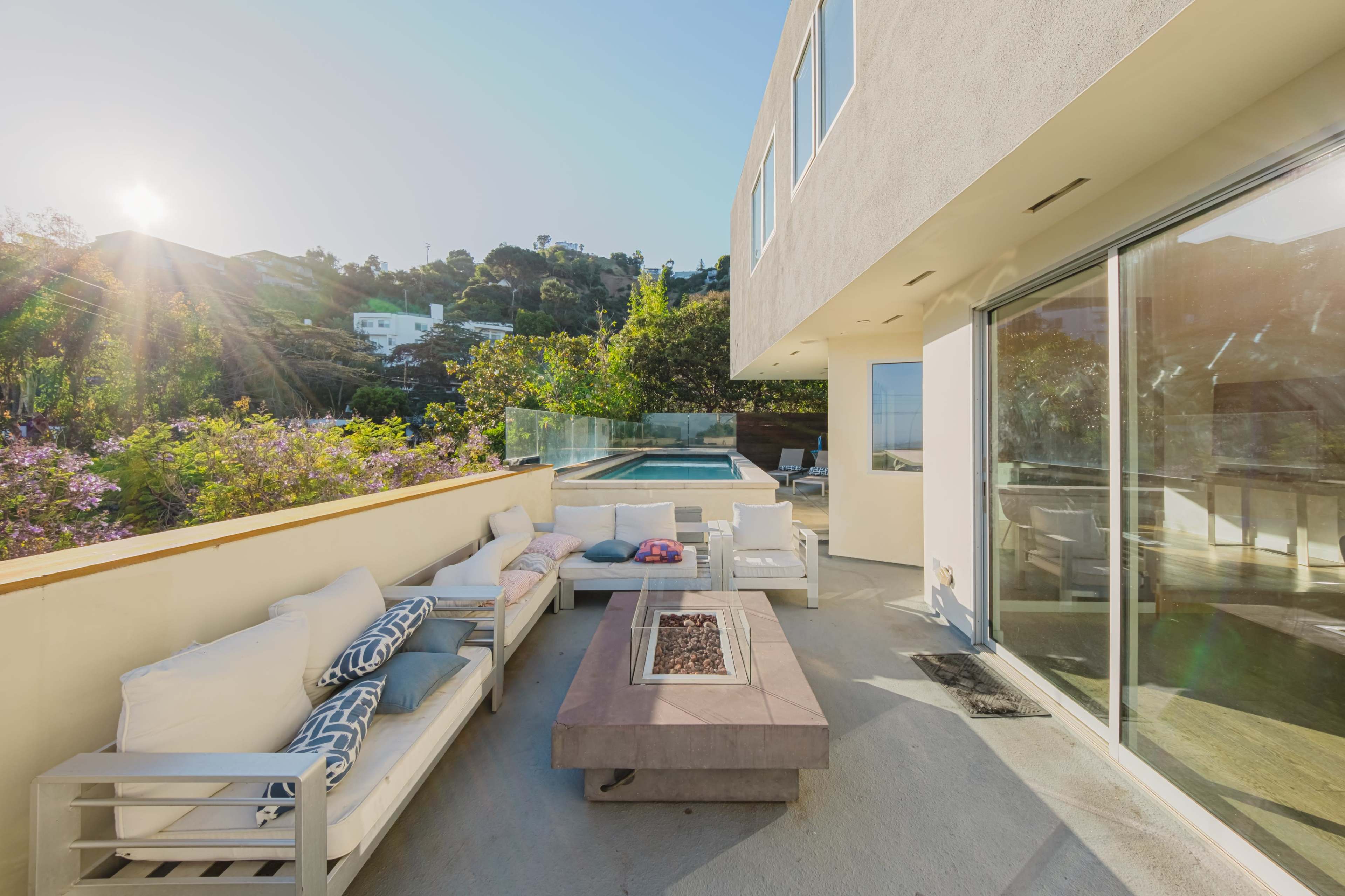 A modern patio features a seating area with white couches surrounding a rectangular fire pit, overlooking a pool and hillside greenery.